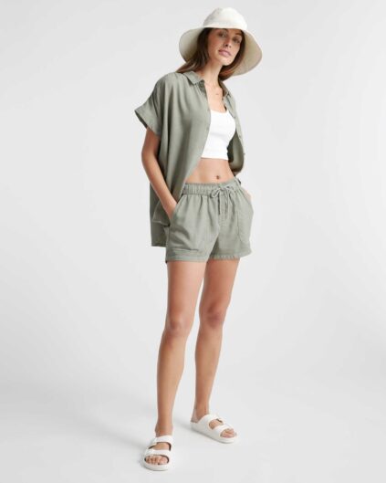 A woman wearing a pale green short-sleeve button-up shirt, matching shorts, a white crop top, white sandals, and a wide-brim hat stands against a plain background.