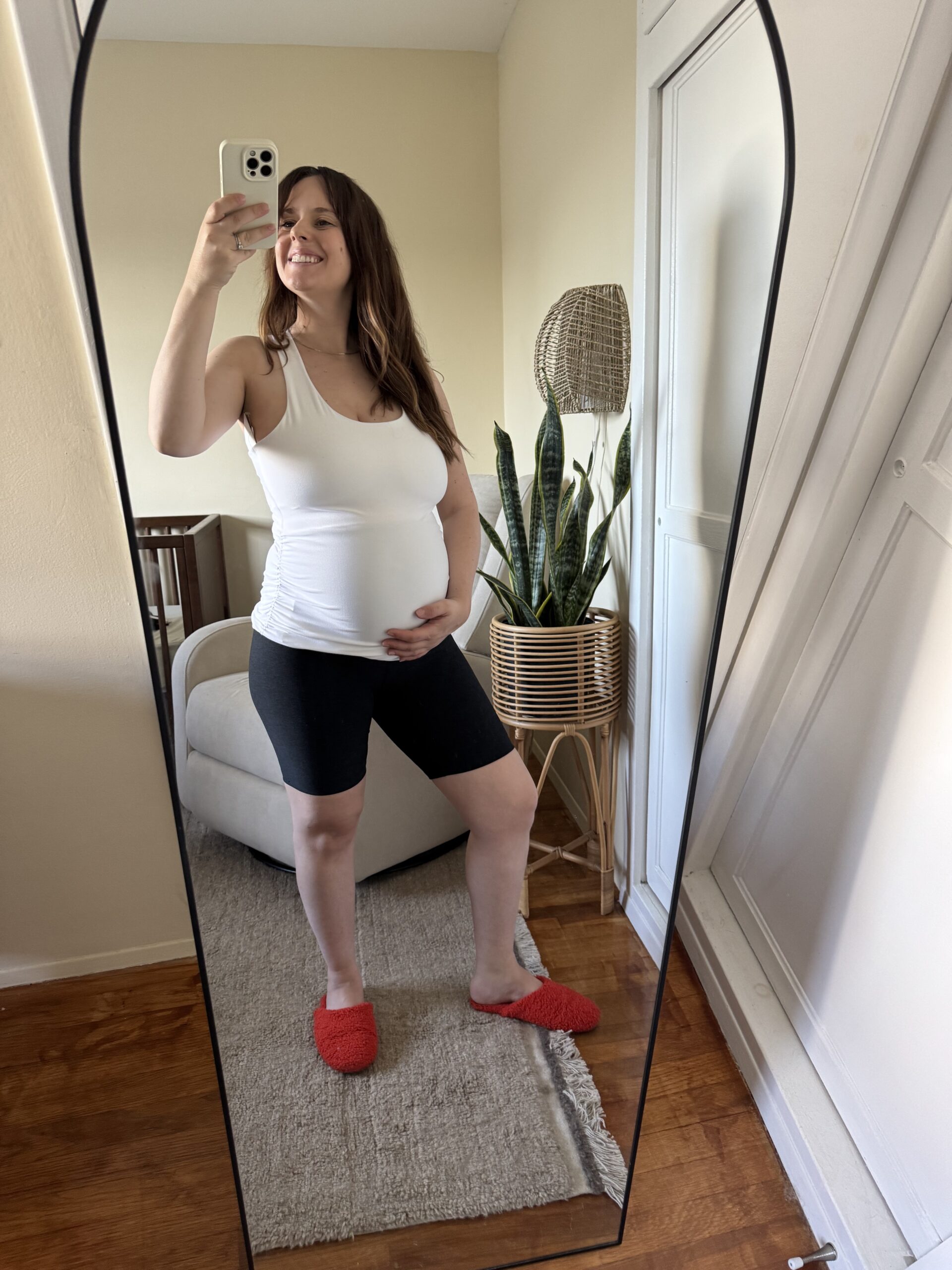 A woman wearing a white tank top, black shorts, and red slippers takes a mirror selfie, holding her pregnant belly in a cozy room with plants and neutral decor.