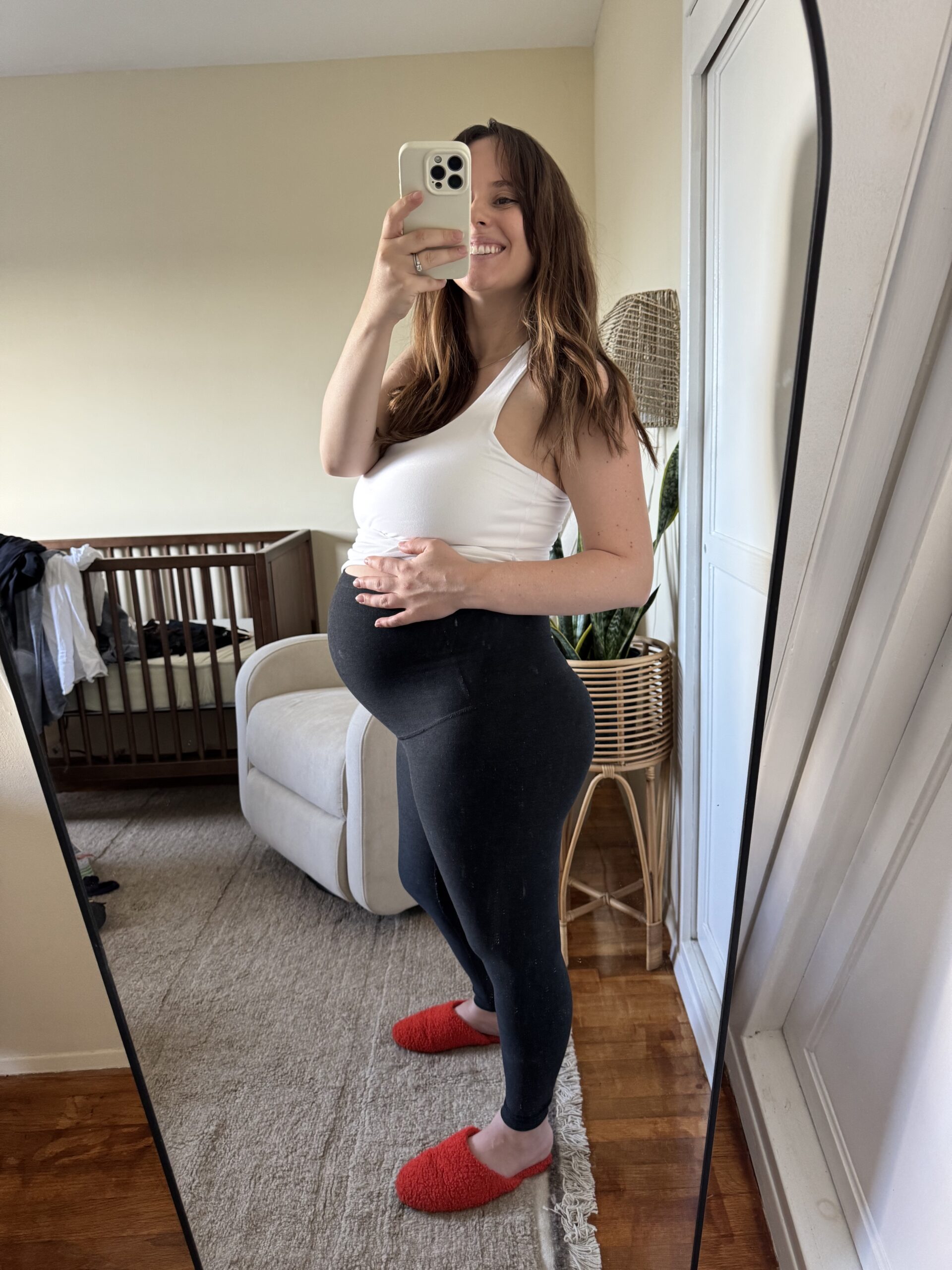 A woman takes a mirror selfie in a bedroom, wearing a white top, black leggings, and red slippers, holding her pregnant belly. A crib and chair are visible in the background.