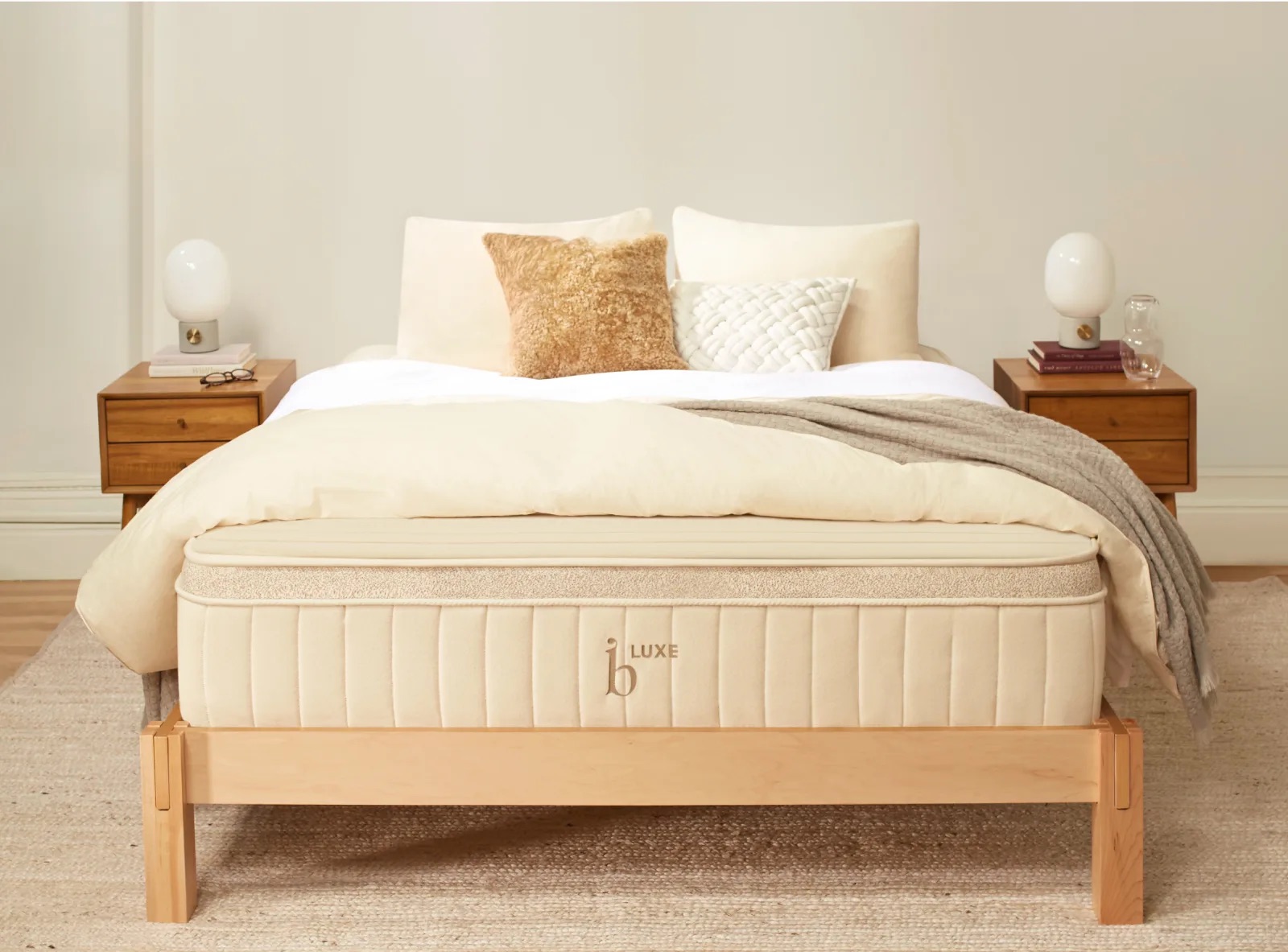 A neatly made bed with a beige mattress labeled "Luxe" is flanked by two wooden nightstands. Each nightstand has a lamp. The bed has light-colored bedding and several decorative pillows.