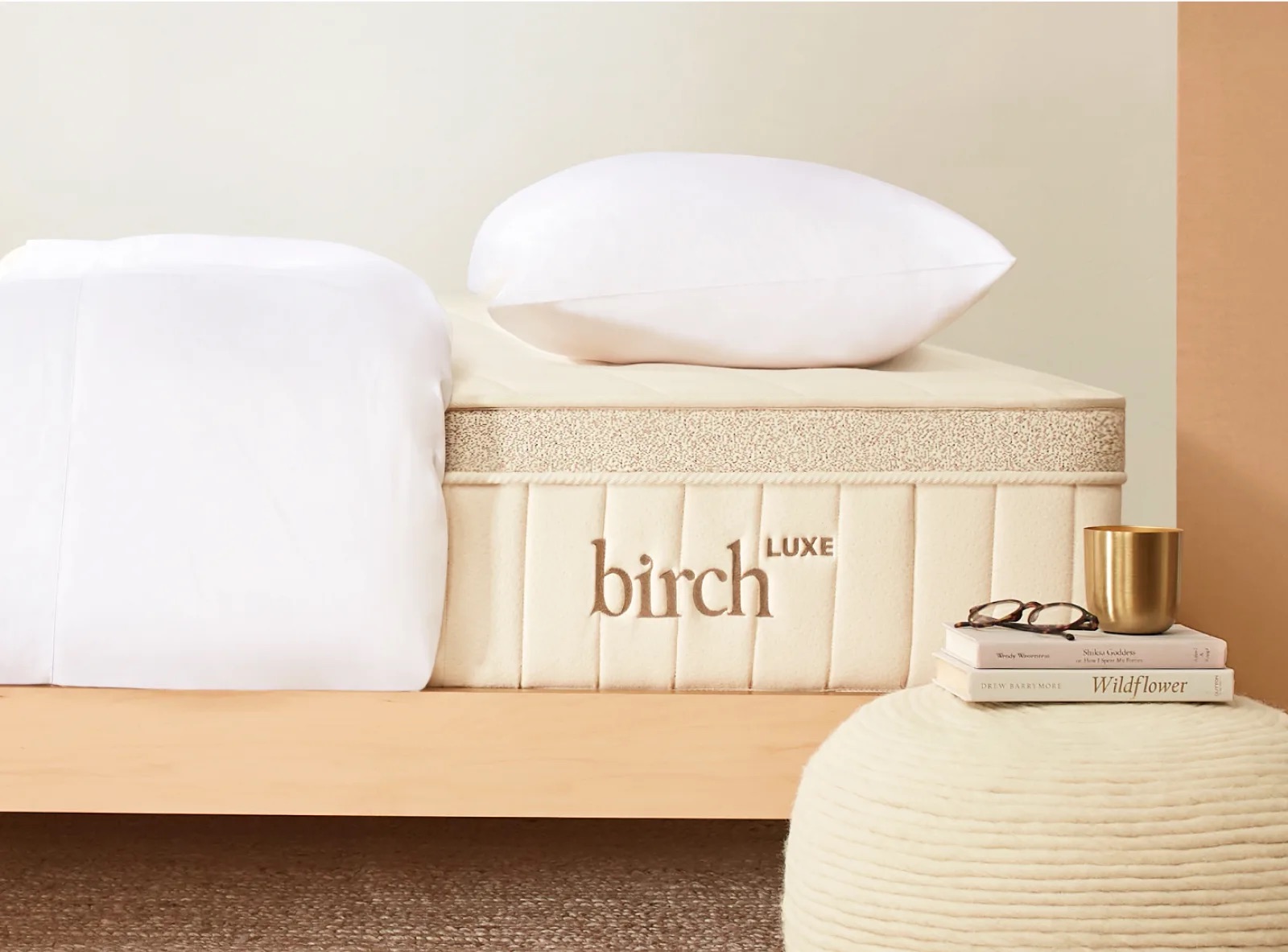 Mattress with "Birch Luxe" embroidered, topped with white bedding and a pillow. Nearby are a white pouf, a stack of books, and a gold cup.