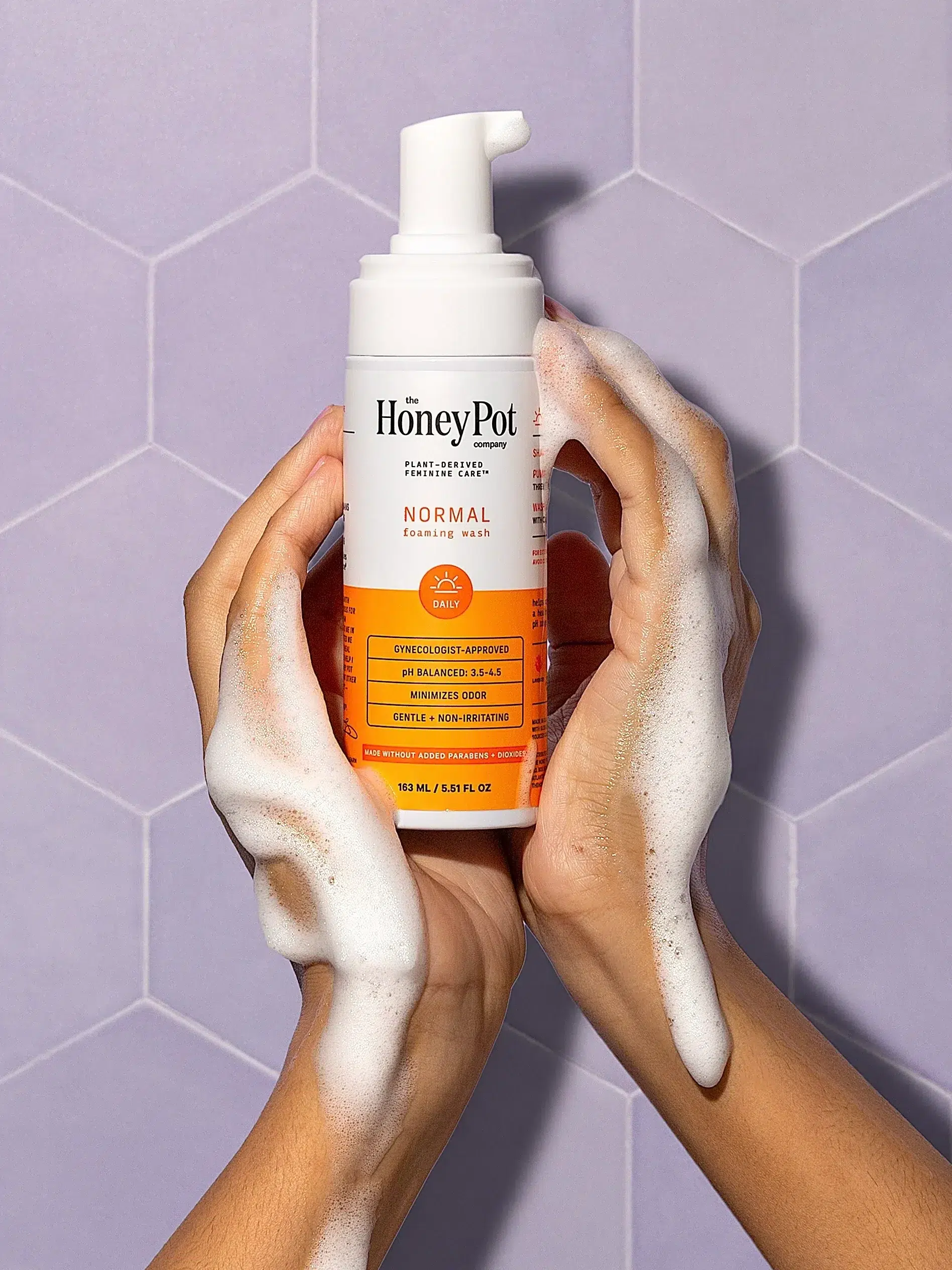 Hands holding a bottle of The Honey Pot Company's normal foaming wash against a hexagonal tiled background, with soap suds visible.