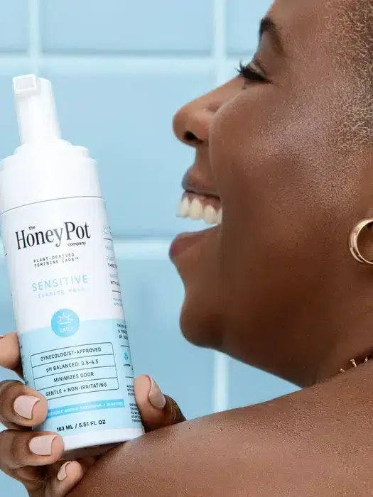 Person smiling while holding a bottle of Honey Pot Sensitive Foaming Wash in front of a blue tiled background.