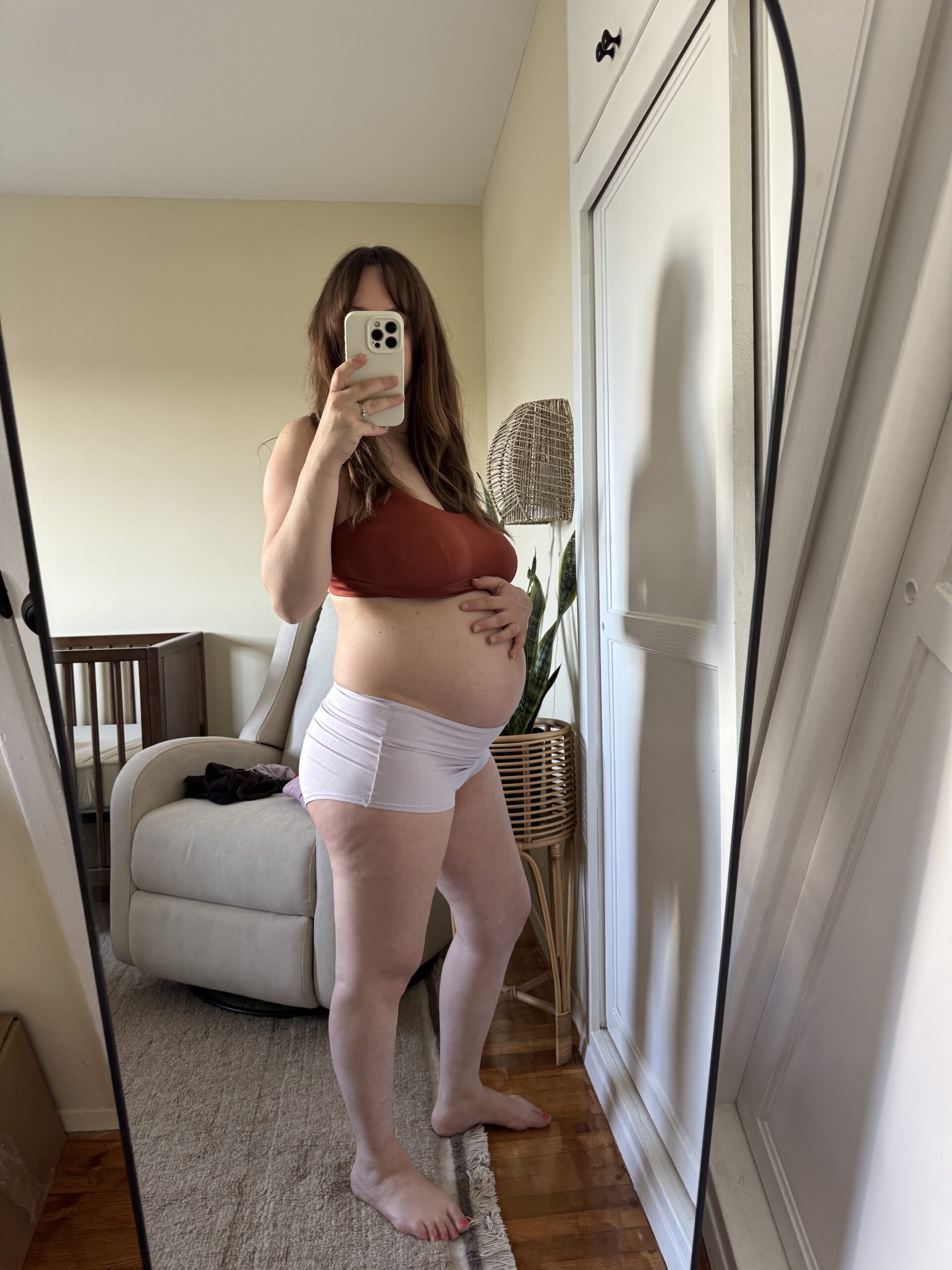A person takes a mirror selfie in a bedroom, showing a baby bump. They are wearing a rust-colored top and white shorts, with one hand on their stomach.
