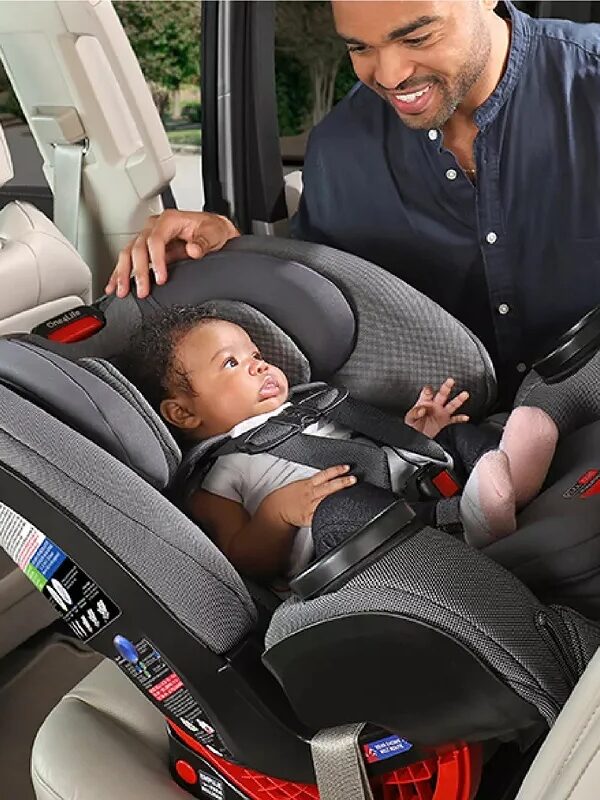 An adult secures a baby in a car seat inside a vehicle. The baby is facing forward, and both appear content.