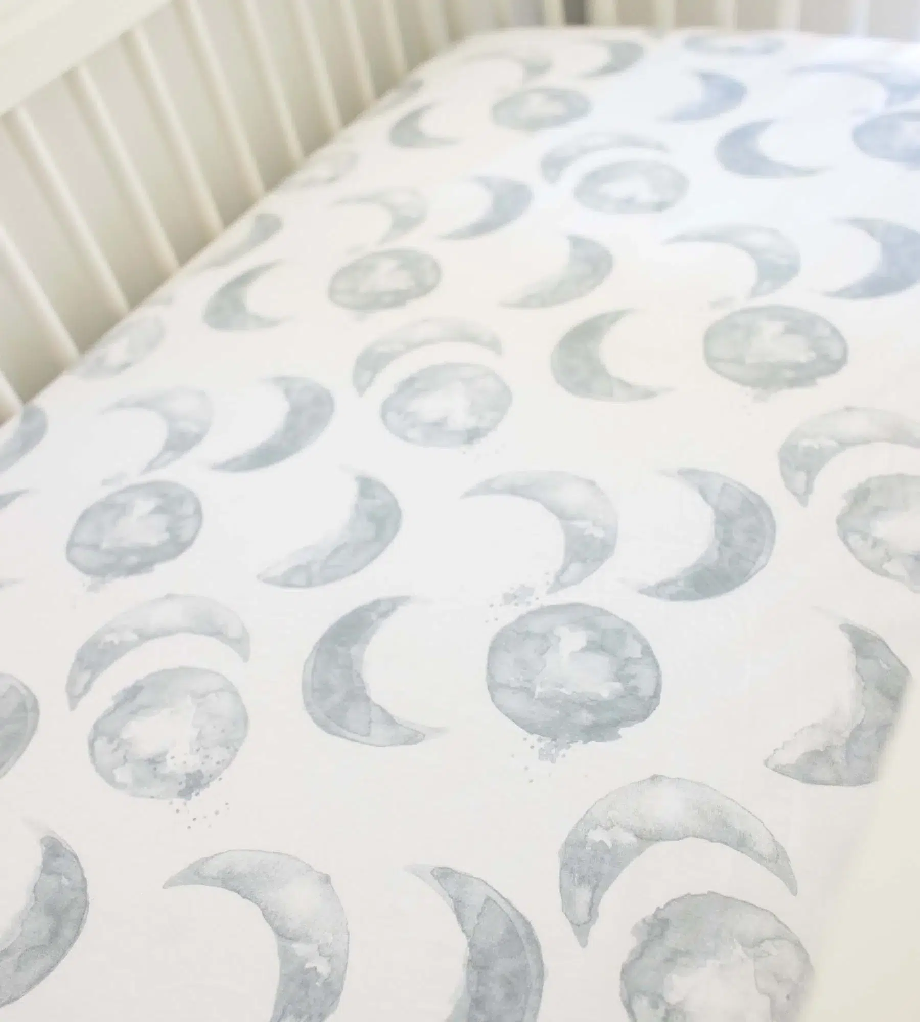 A crib mattress with a fitted sheet featuring a repeating pattern of watercolor-style crescent moons and full moons in soft gray tones.