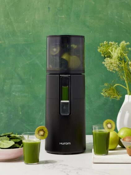 A black Hurom juicer with green juice inside, surrounded by a glass of green juice, spinach, kiwis, and a plant, against a green textured background.
