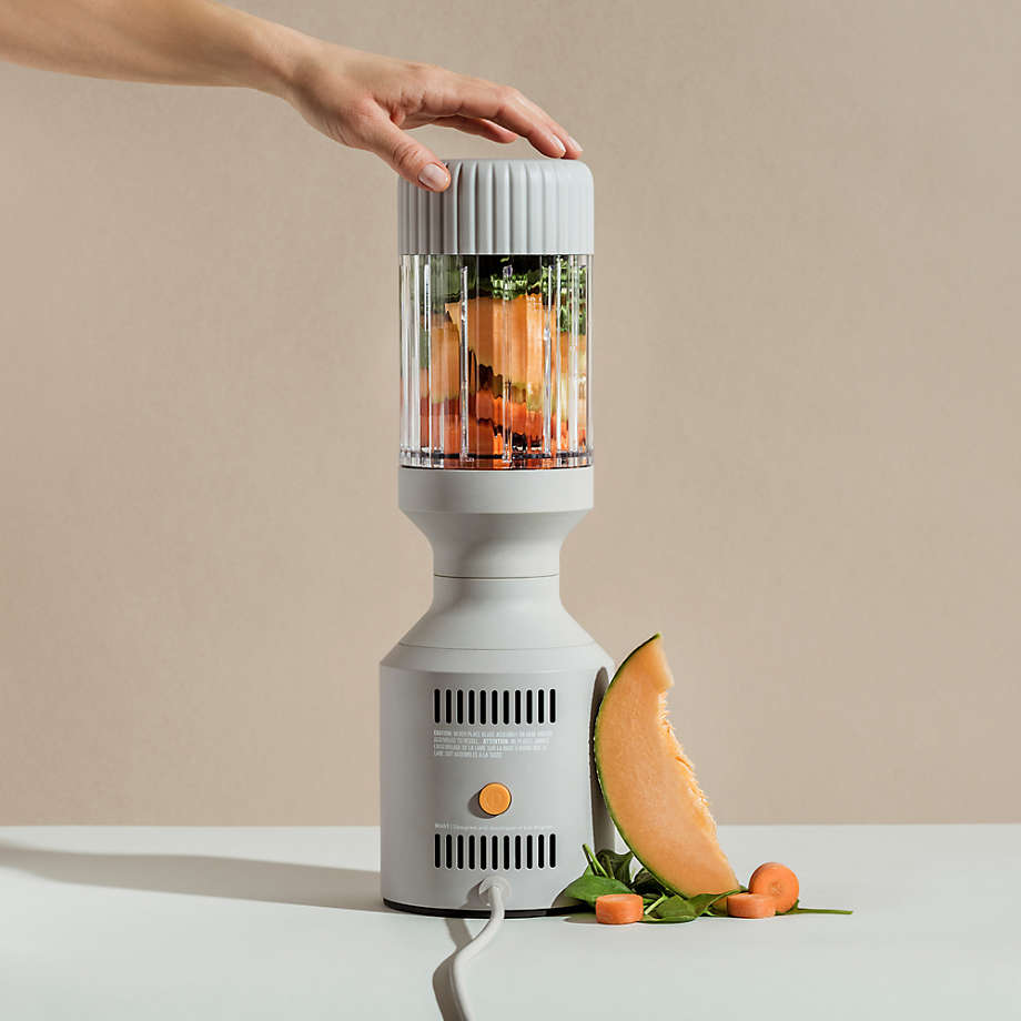 Hand pressing a button on a vertical blender with fruits and greens inside. A slice of cantaloupe and some spinach leaves are on the table next to the blender.