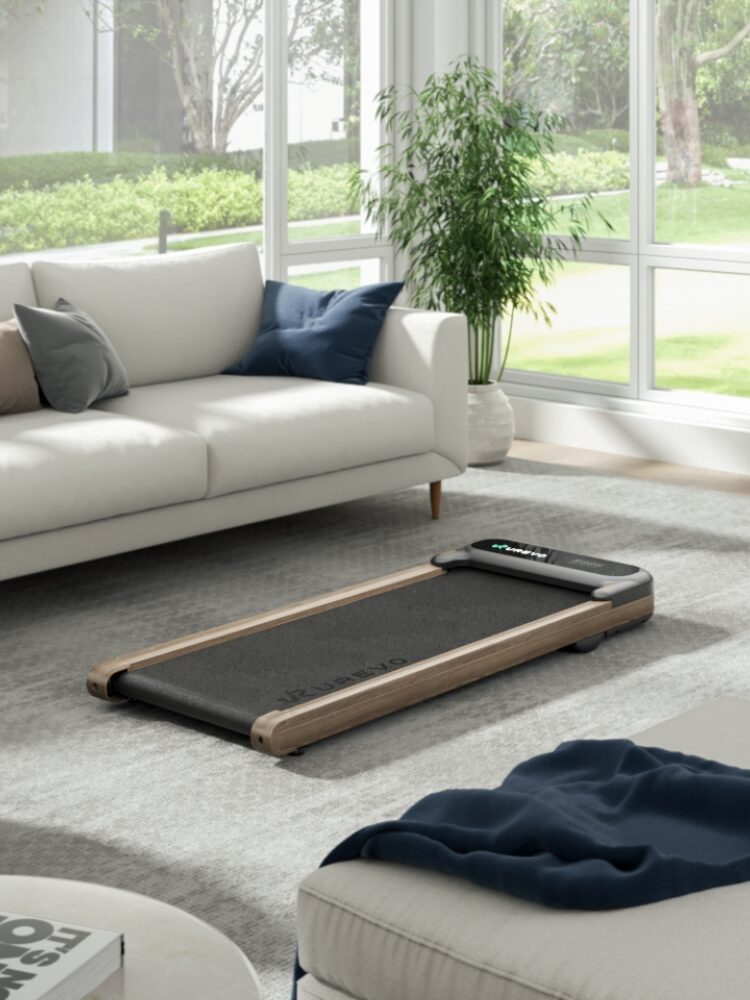 A compact treadmill sits on a gray rug in a modern living room with a white couch, a blue blanket, and large windows showing greenery outside.