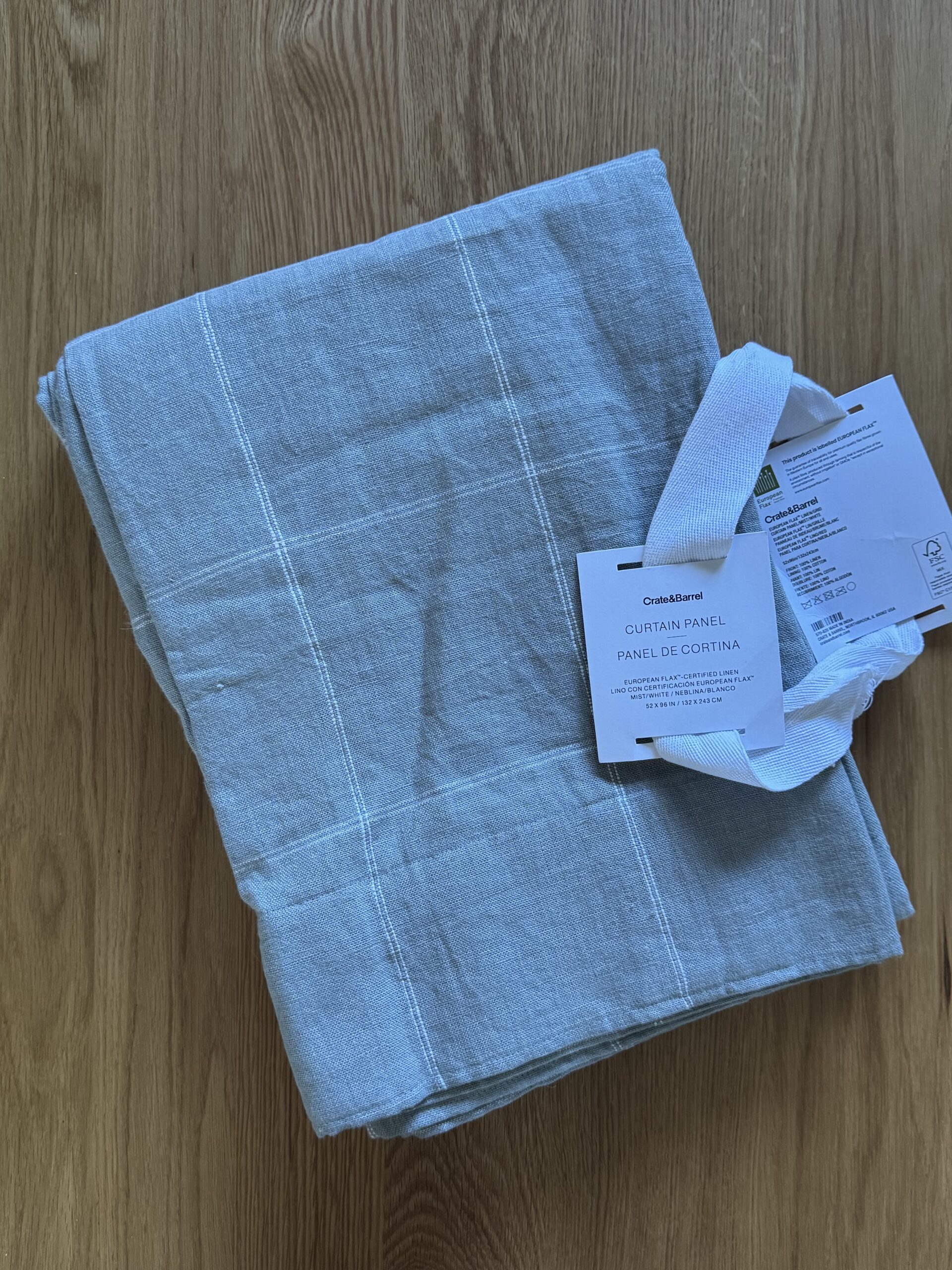 Folded curtain panel with a grid pattern rests on a wooden surface. A white tag is attached, displaying product information.