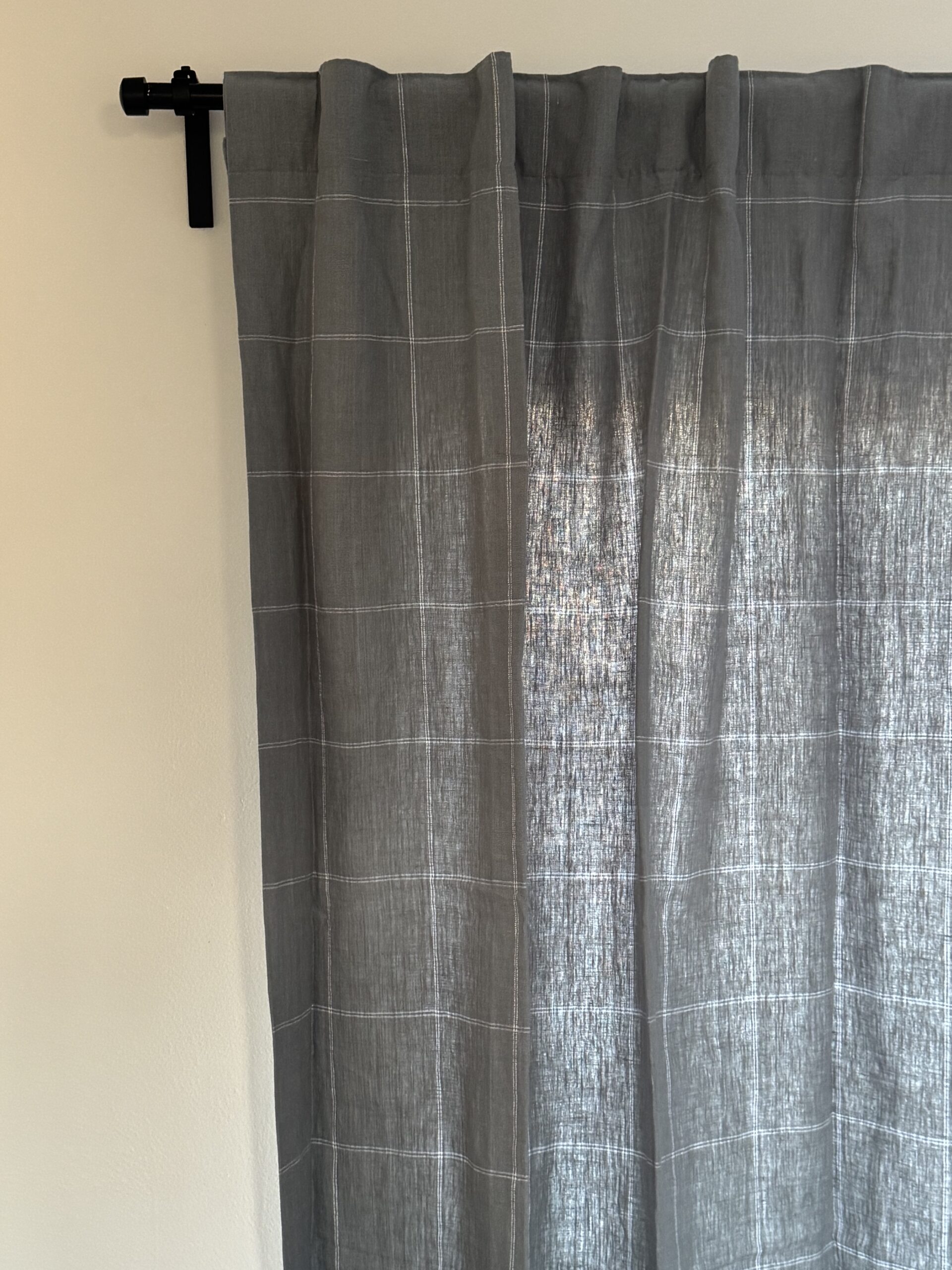Grey curtain with a subtle checkered pattern on a black rod, partially drawn to the left against a light wall.