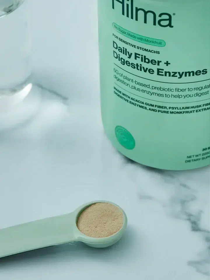 A green container labeled "Daily Fiber + Digestive Enzymes" next to a glass of water and a green measuring spoon filled with powder on a marble surface.