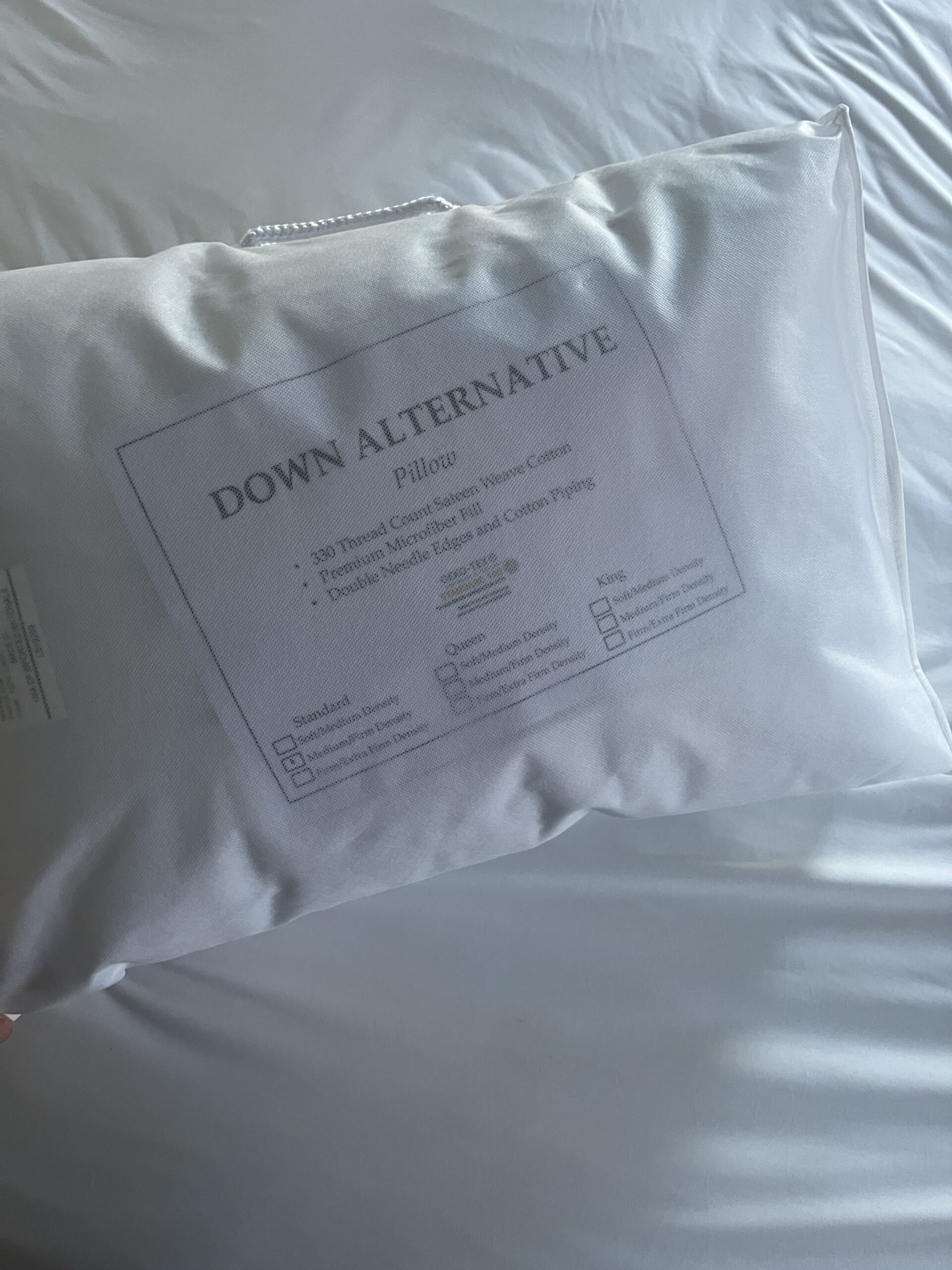 A white "Down Alternative" pillow on a bed, featuring a label detailing its 300 thread count, micro sateen cotton, and double-needle edge with piping.