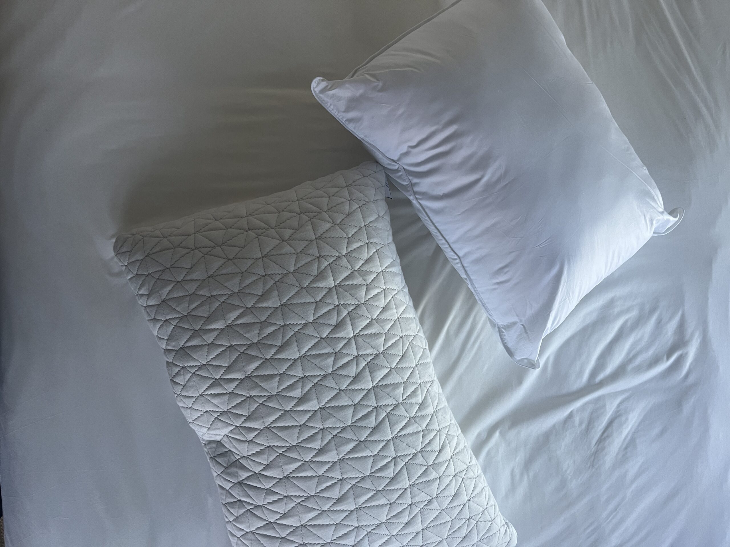 Two white pillows on a white bed: one with a quilted pattern and the other plain.