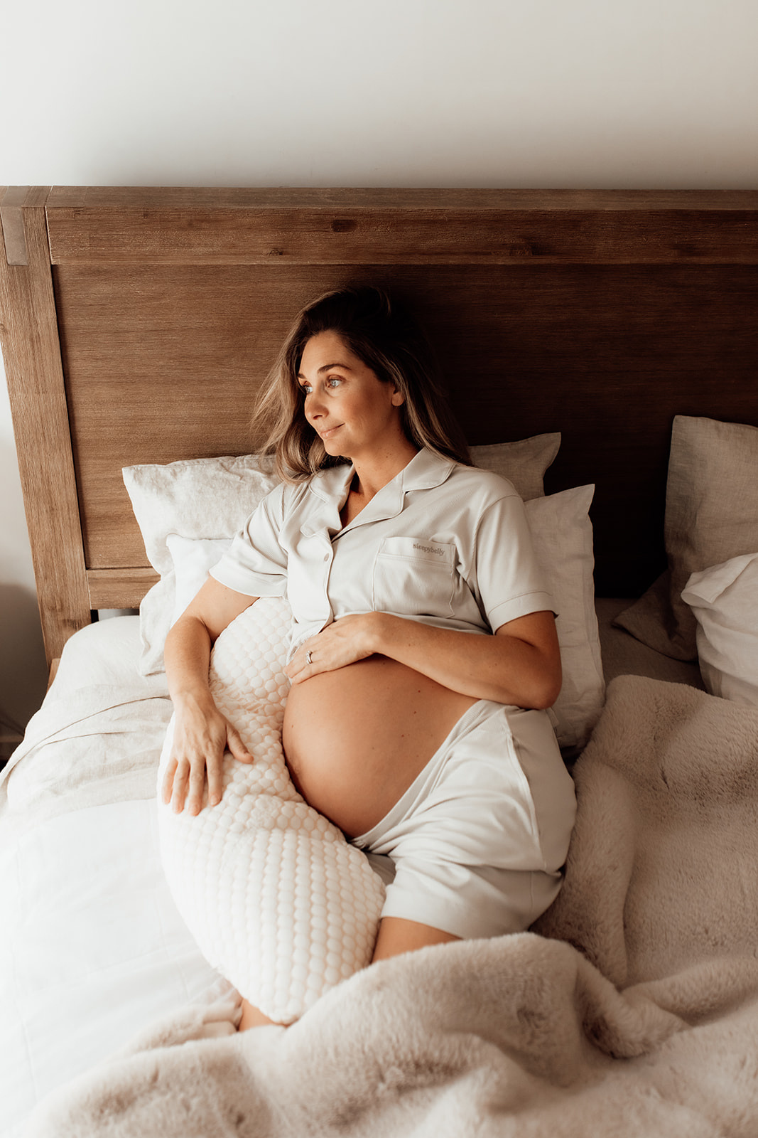 Pregnant woman in pajamas reclines on a bed, resting a pillow on her belly.