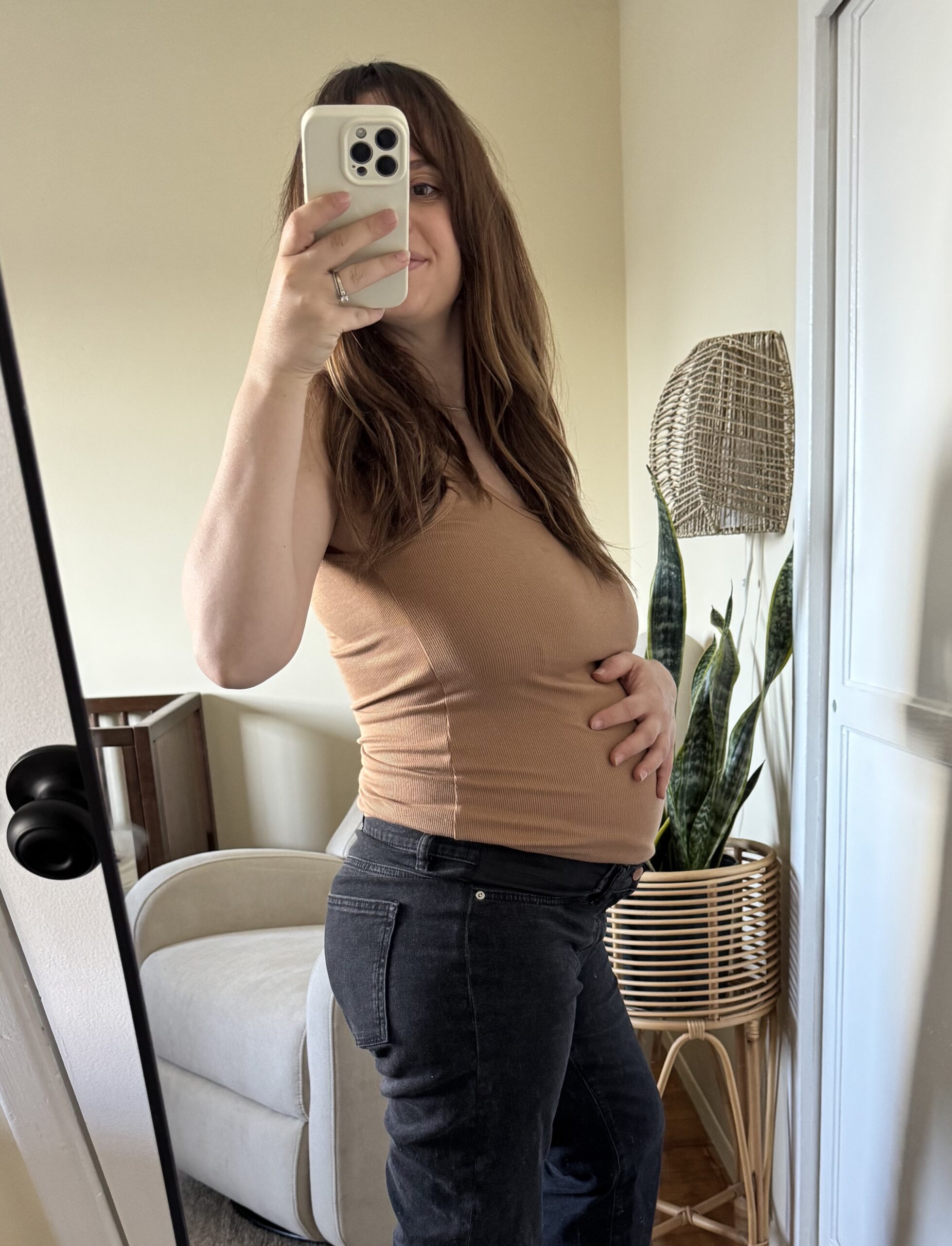 A woman takes a mirror selfie, holding her phone in one hand and her baby bump with the other, standing in a room with a chair and a potted plant in the background.