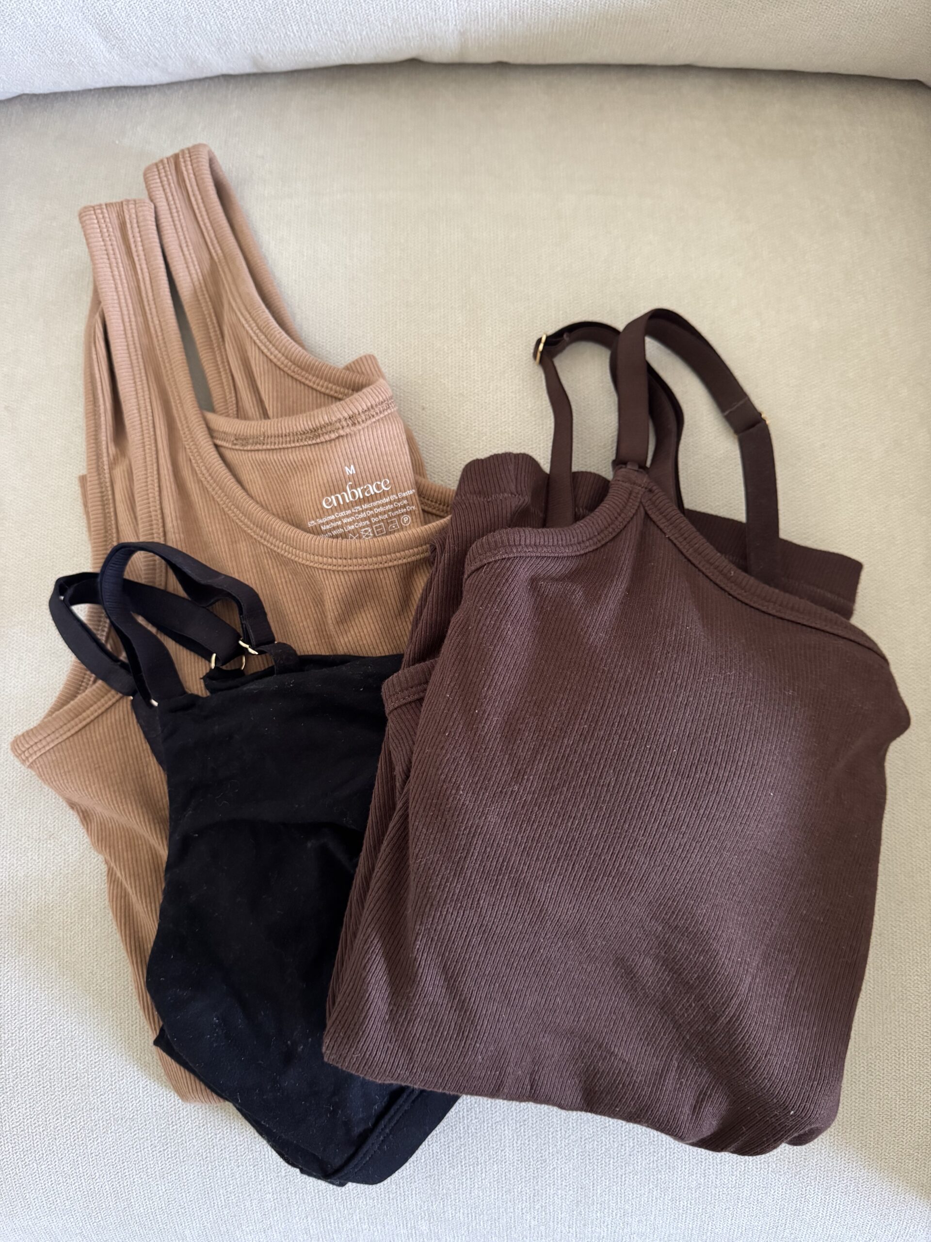 Three ribbed tank tops in beige, black, and brown laid out on a light-colored fabric surface.