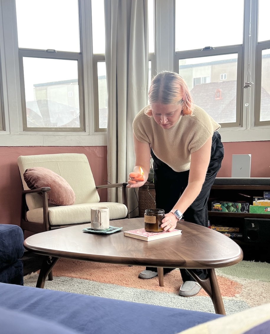 A person lights a candle on a wooden coffee table in a living room with large windows, an armchair, and a shelf in the background.