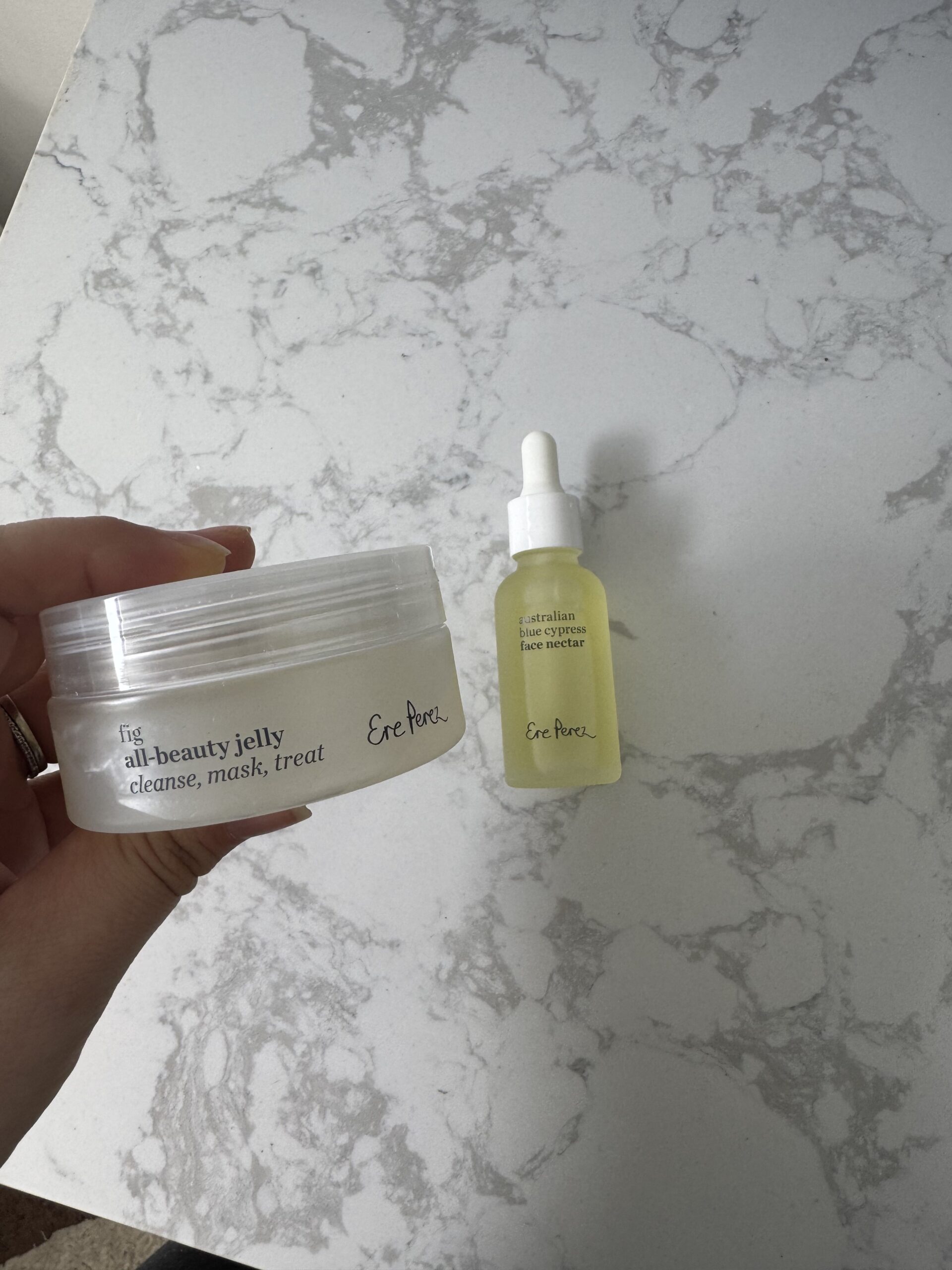 Hand holding a jar of fit all-beauty jelly next to a dropper bottle labeled Australian face nectar by Ere Perez, on a marble surface.