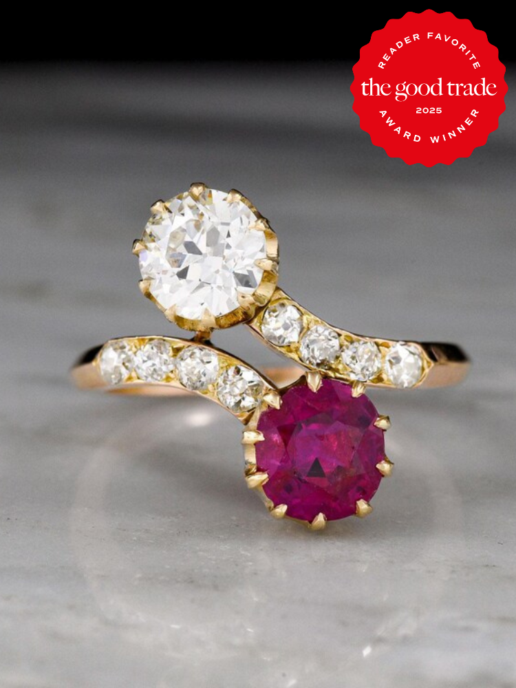 Gold ring with a large clear and red gemstone, each surrounded by smaller clear stones, set against a marble background. Award badge in the top corner.