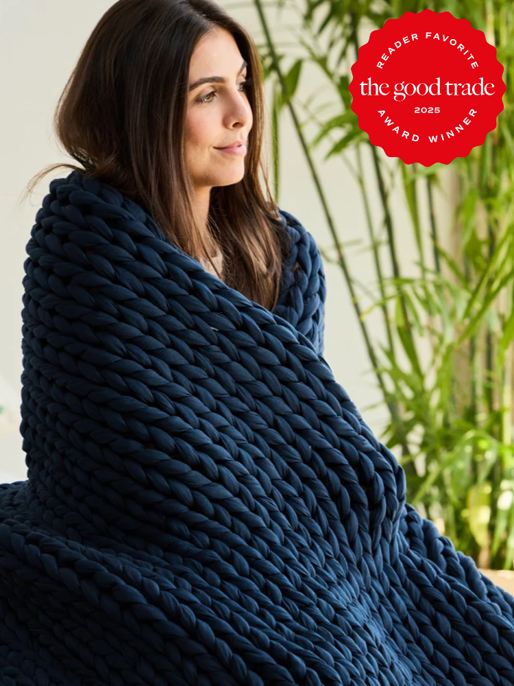 Woman wrapped in a thick, dark blue chunky knit blanket sits indoors near green plants; "The Good Trade 2025 Award Winner" badge is in the top right corner.