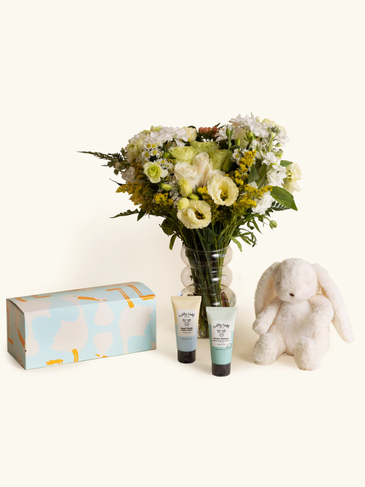 A bouquet of white and yellow flowers in a glass vase, two tubes of lotion, a light blue gift box, and a white plush bunny arranged on a cream background.