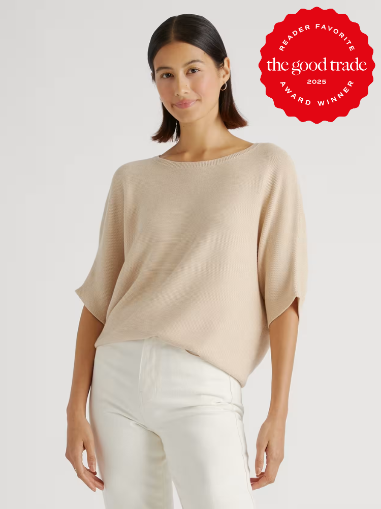 A woman wearing a light beige short-sleeve sweater and white pants stands against a plain background. A red "The Good Trade Award Winner 2025" badge appears in the top right corner.