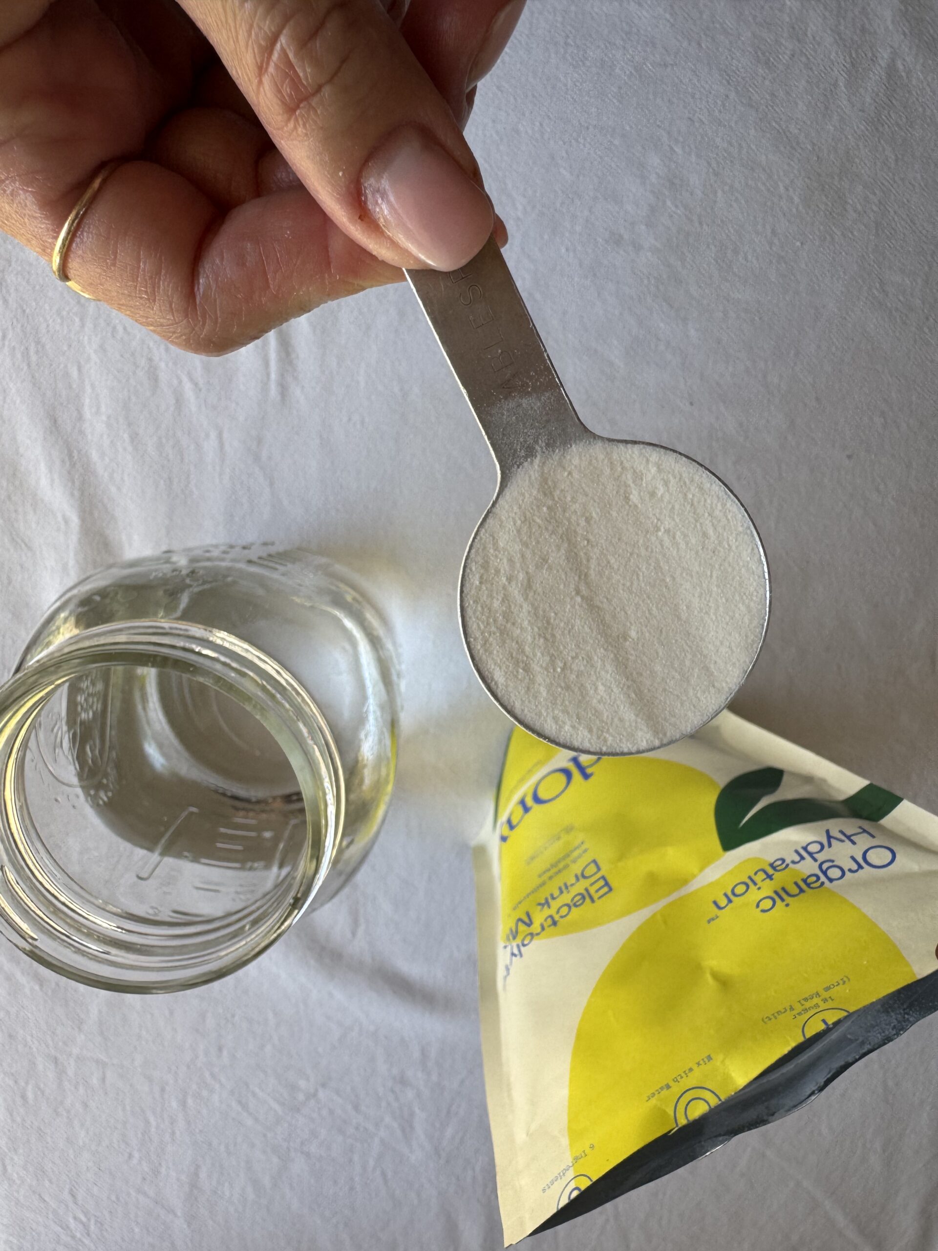 A hand holds a metal tablespoon of white powder above a glass jar of water and a yellow and white bag labeled "organic hydration.