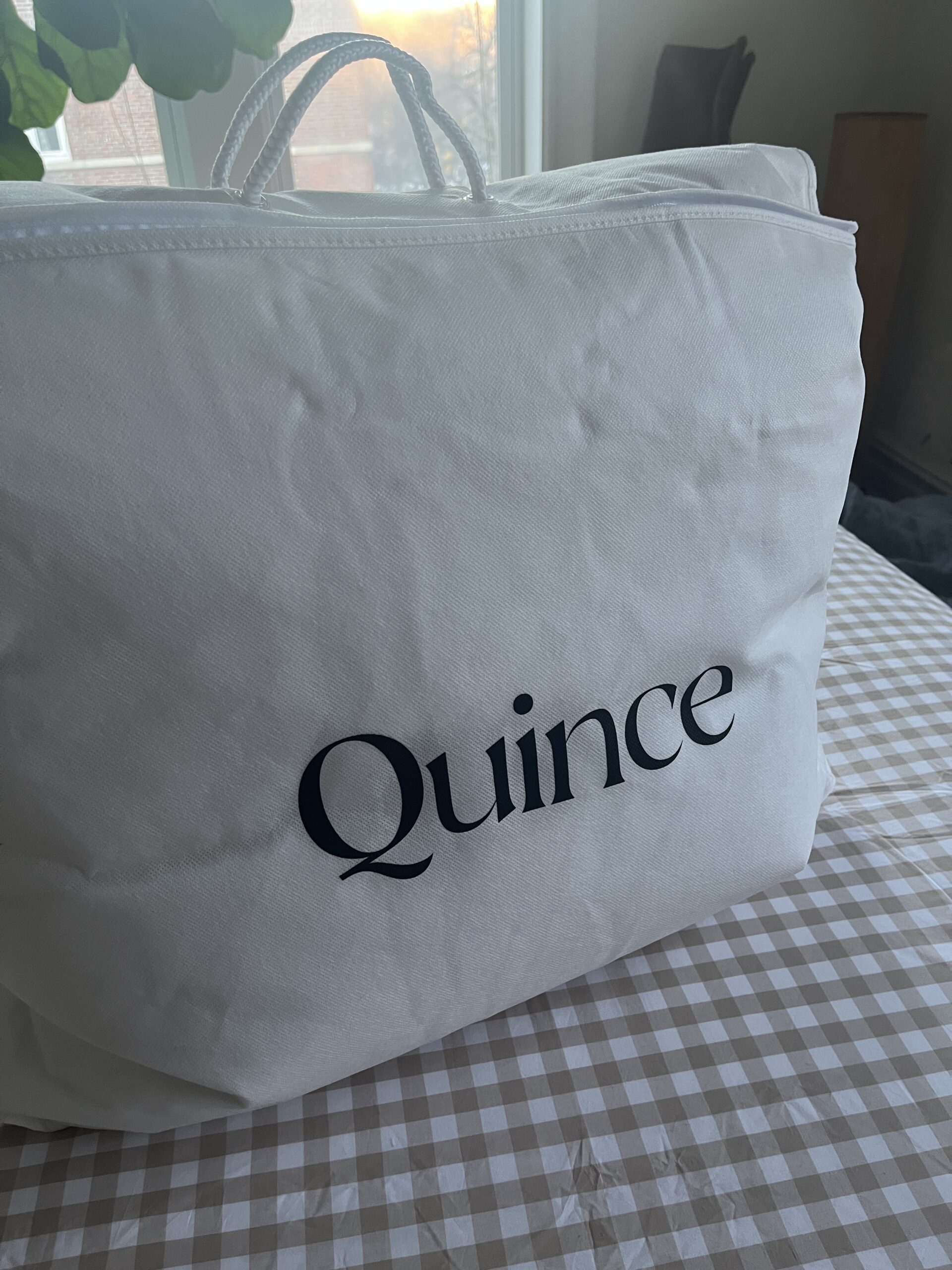 A white bag with handles on a checkered table, labeled "Quince" in large black text.