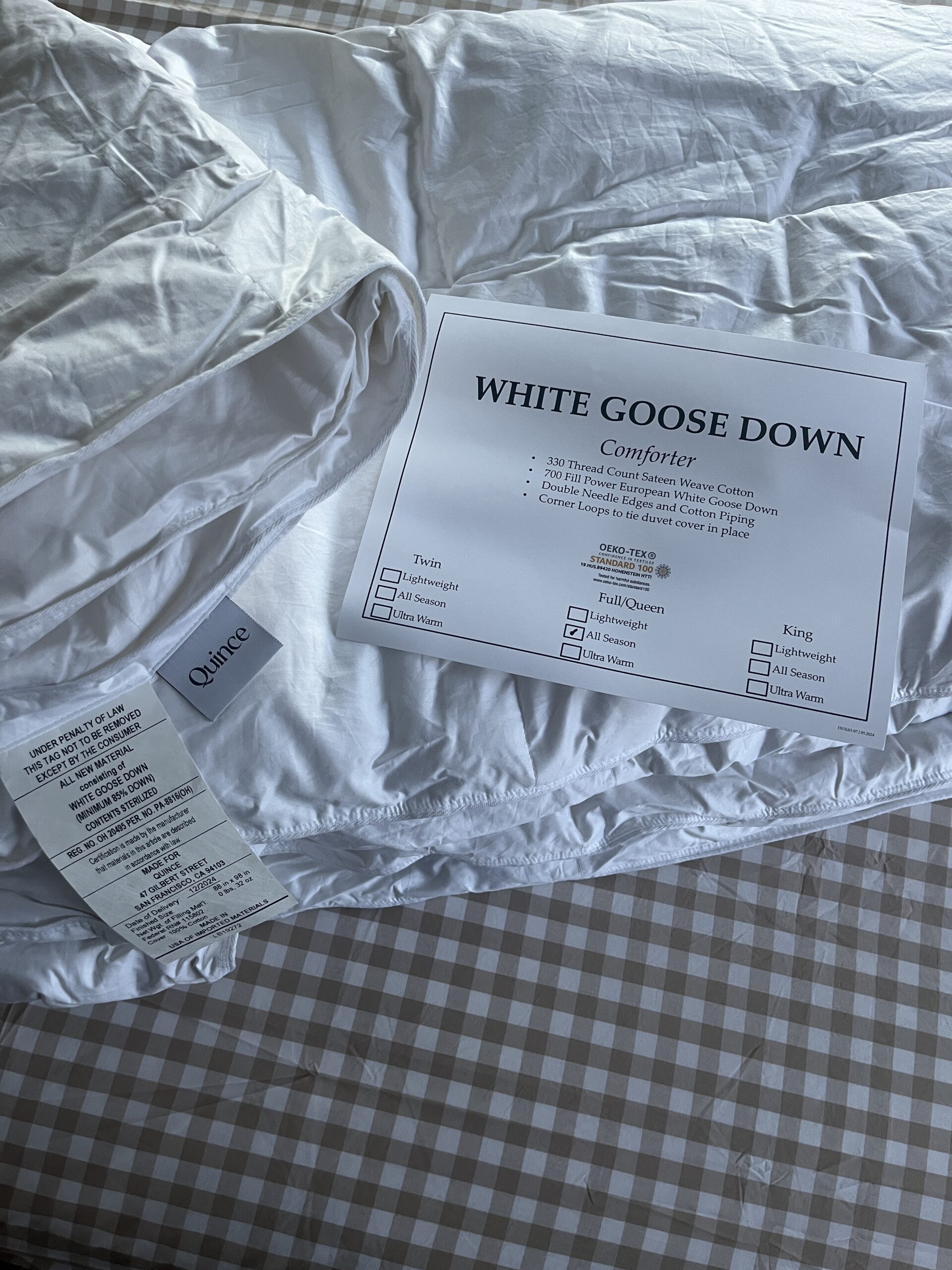 White goose down comforter on a checkered bedspread with a label detailing size, care instructions, and features.