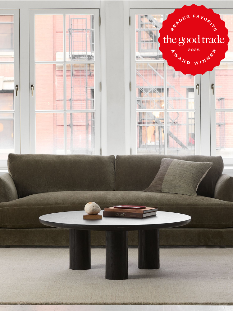 A modern living room with a green sofa, round coffee table, and book, set against large windows. A "Reader Favorite" award badge from "The Good Trade" for 2023 is in the top right corner.