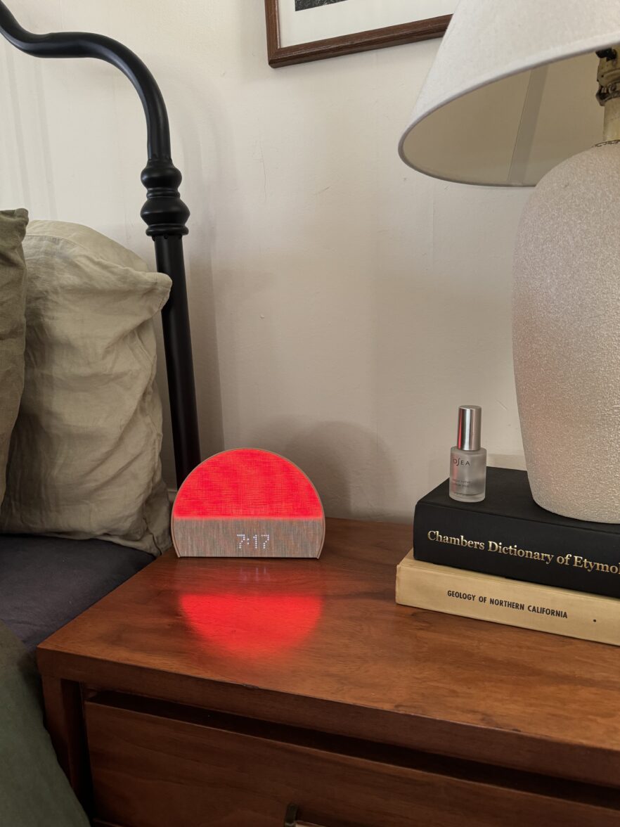 A red-lit digital clock reads 7:17 on a wooden bedside table next to books, a lamp, and a small spray bottle, with a bed and pillow nearby.