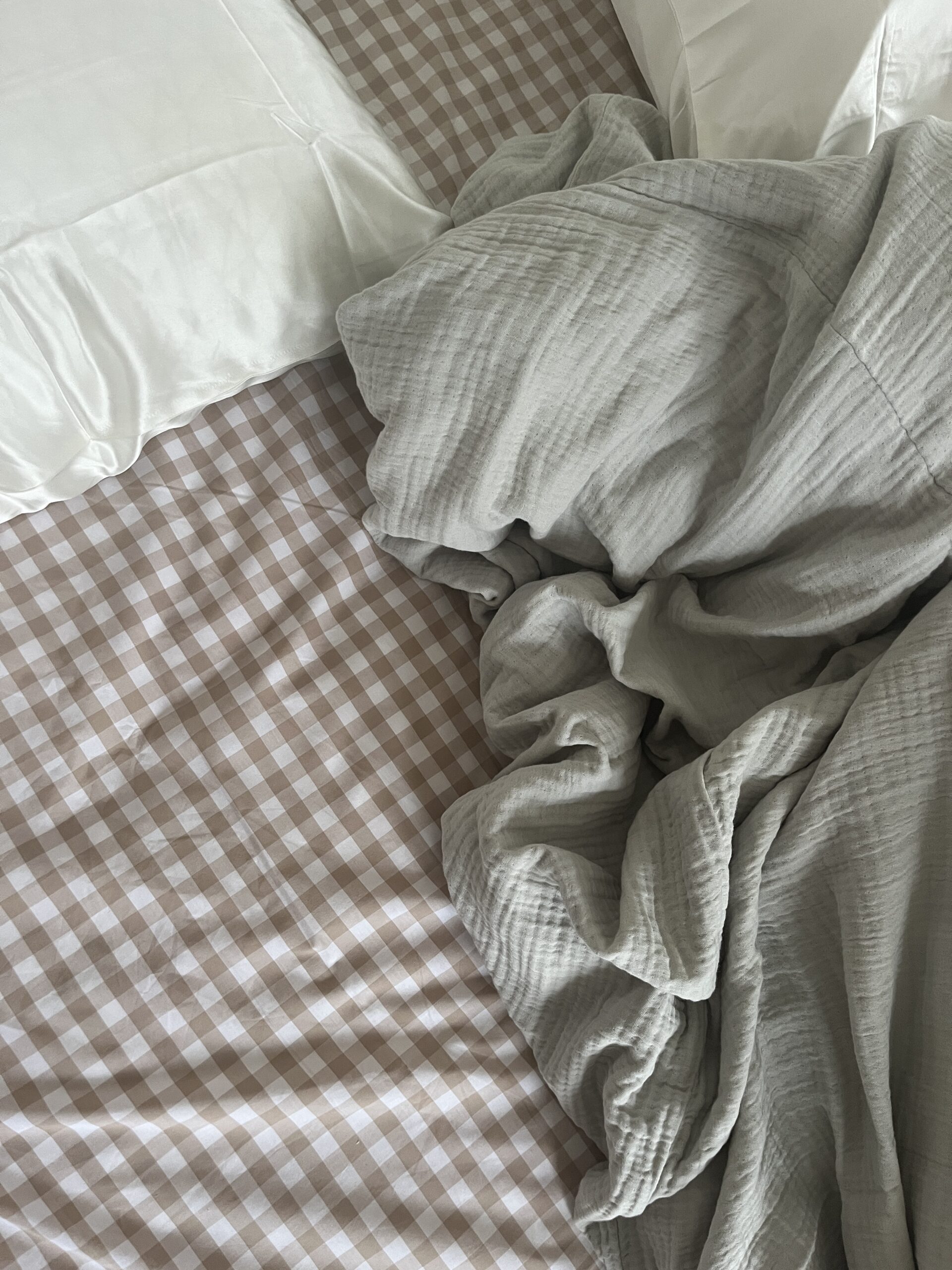 Unmade bed with white pillows and a crumpled gray blanket on a checkered beige and white sheet.
