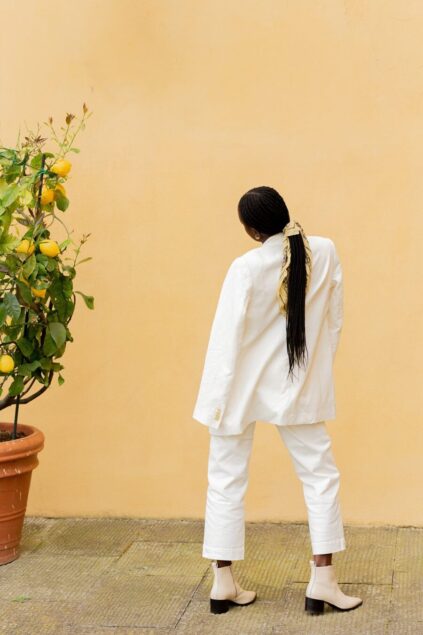Person in white outfit facing a lemon tree against a peach-colored wall, hair tied with a scarf, wearing light-colored boots.