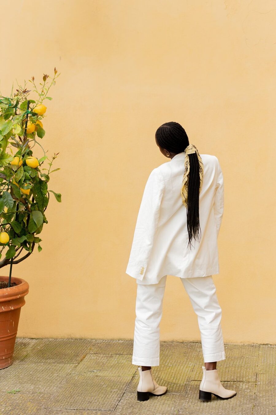 Person in white outfit facing a lemon tree against a peach-colored wall, hair tied with a scarf, wearing light-colored boots.