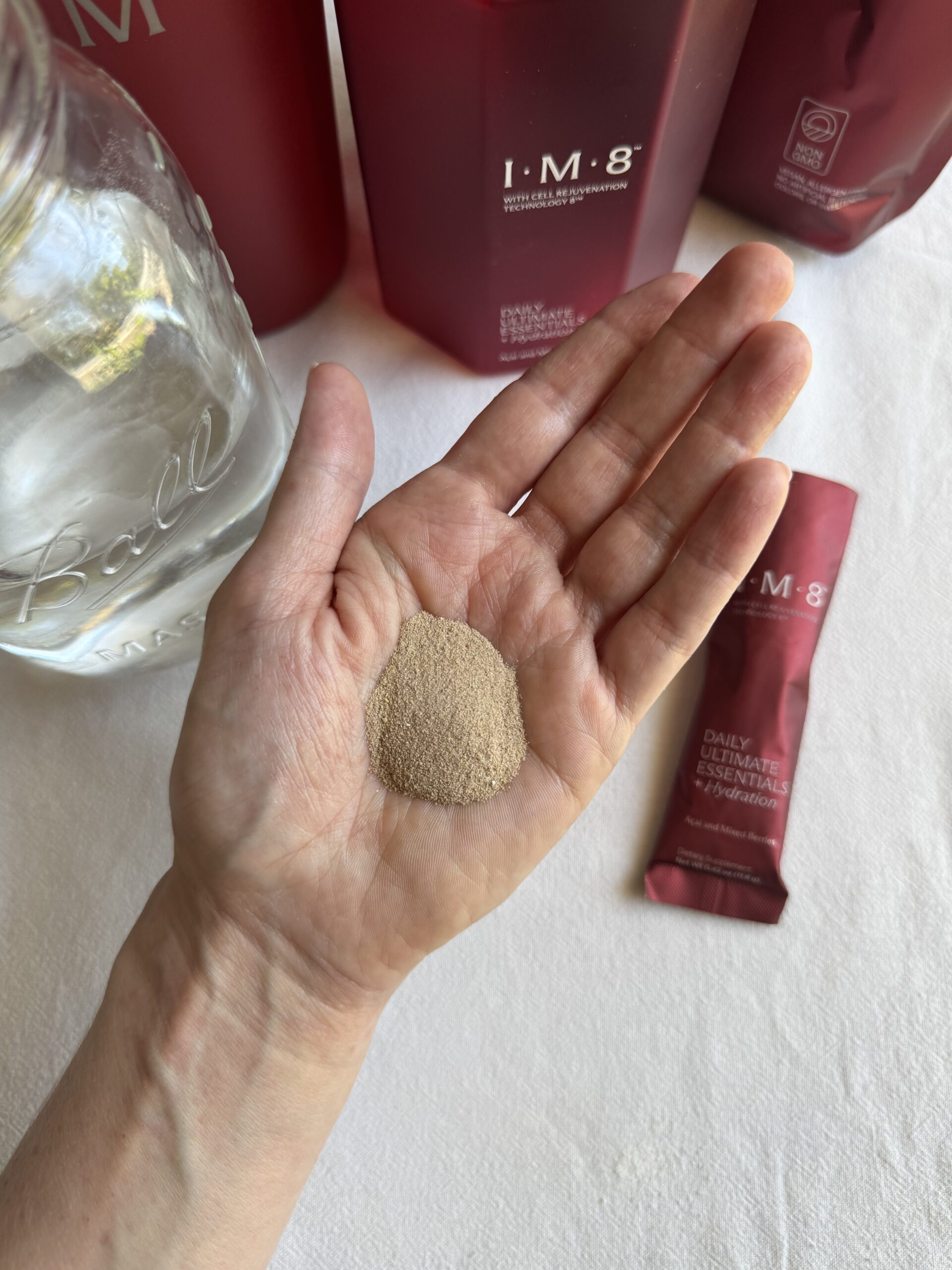 A hand holding a mound of fine beige powder, with a packet labeled "IM-8 Daily Ultimate Essentials" and a glass jar of water in the background.