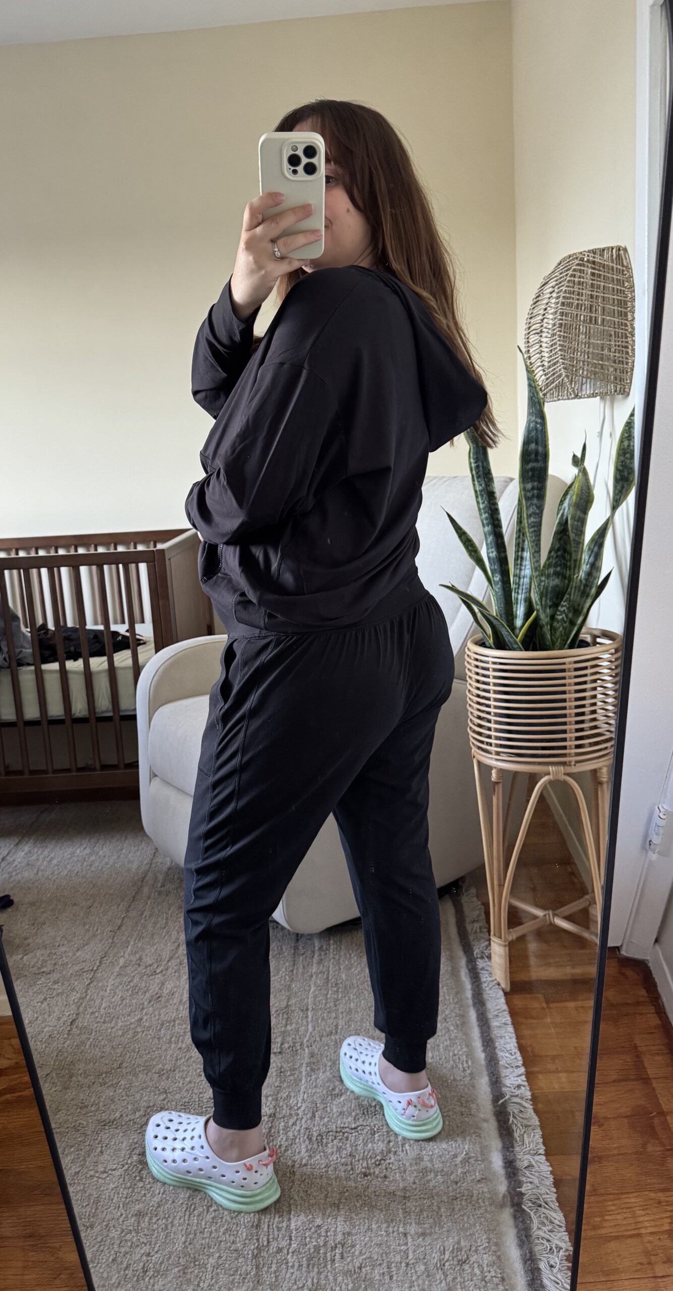 A person in black loungewear and white Crocs takes a mirror selfie in a living room with a crib, a chair, and a large potted plant in the background.