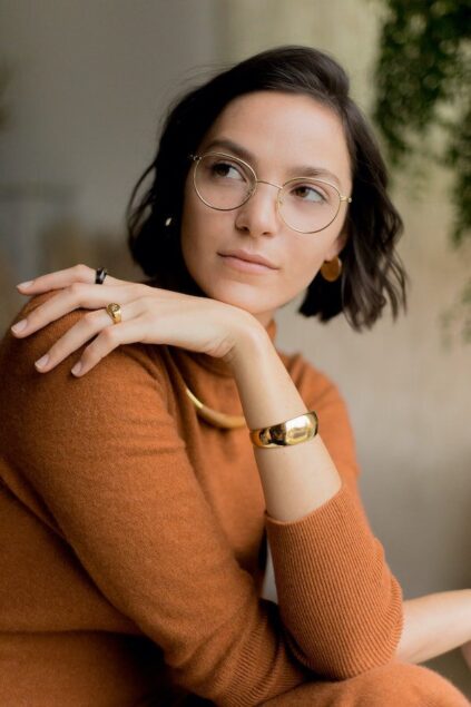 A woman with short brown hair and glasses, wearing an orange sweater and gold jewelry, looks to the side.