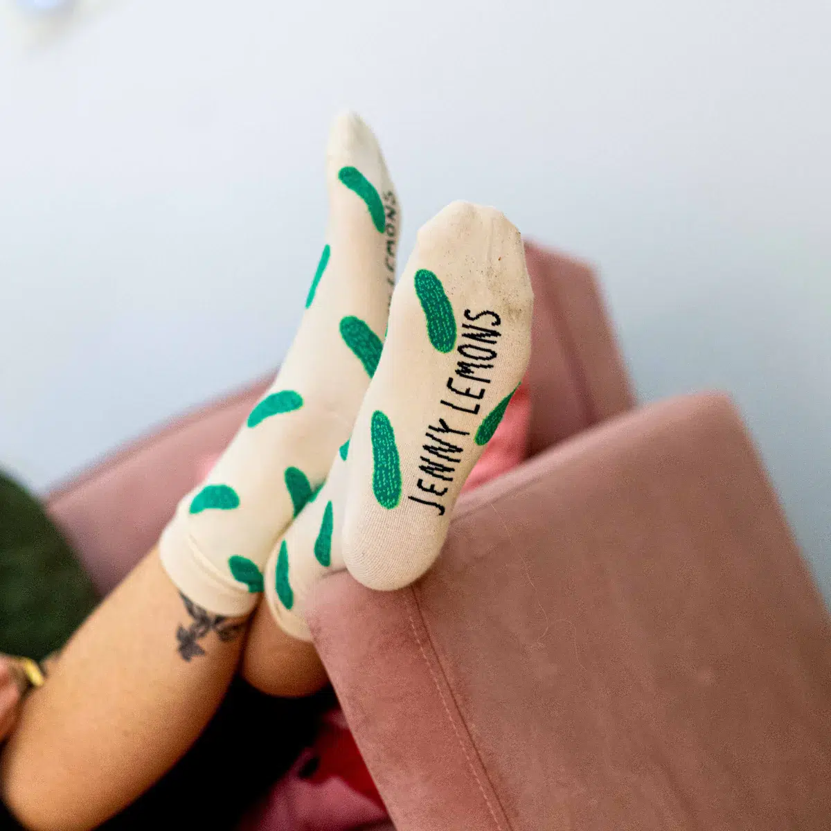 Person resting feet on a pink couch, wearing white organic socks with green pickle patterns and "JENNY LEMONS" written on the soles.