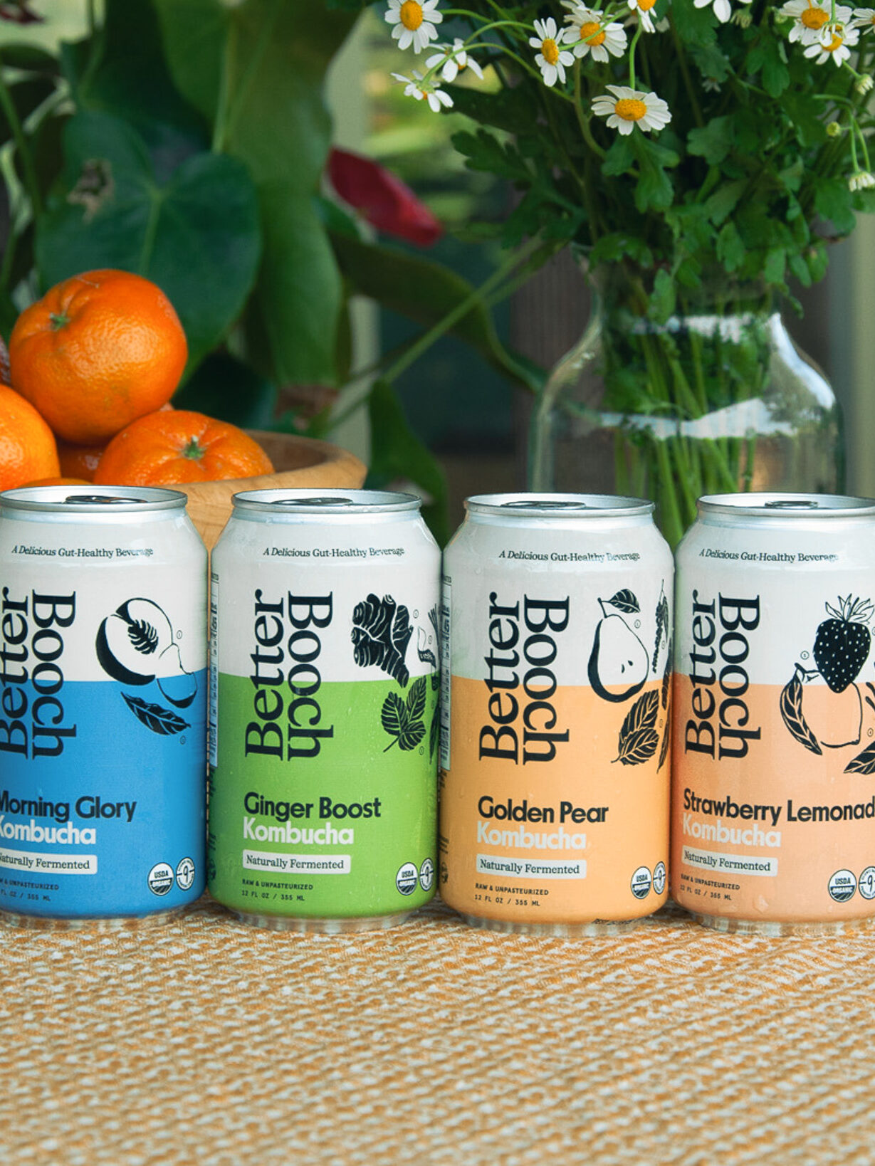 Four cans of Better Booch kombucha in different flavors on a woven surface, with a wooden bowl of oranges and green foliage in the background.