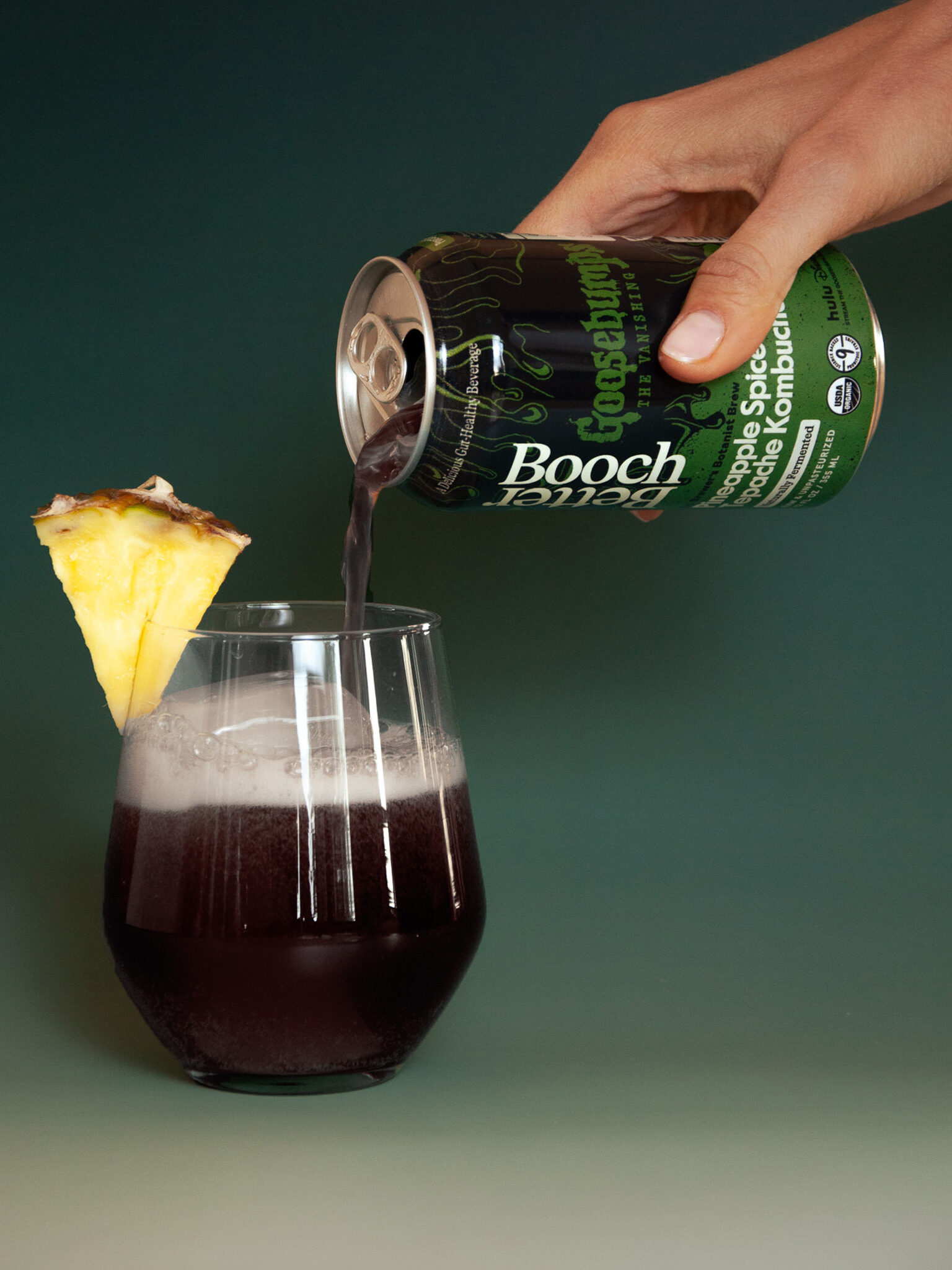 A hand pours a dark liquid from a can labeled "Booch" into a glass with froth at the top. A pineapple slice is garnishing the glass rim.
