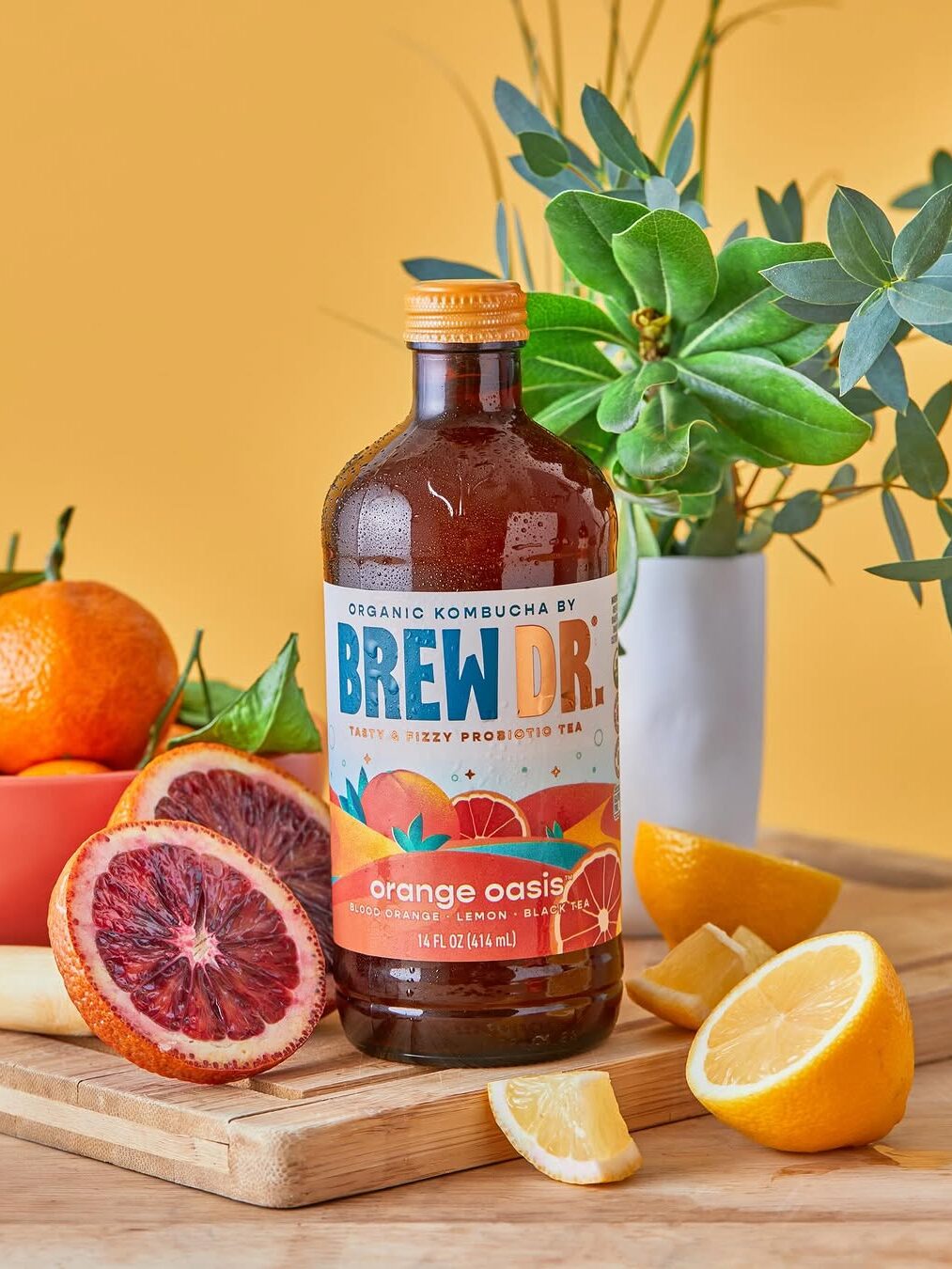 A bottle of Brew Dr. Orange Oasis kombucha on a wooden surface, surrounded by sliced blood oranges, lemons, and green leaves, against a yellow background.