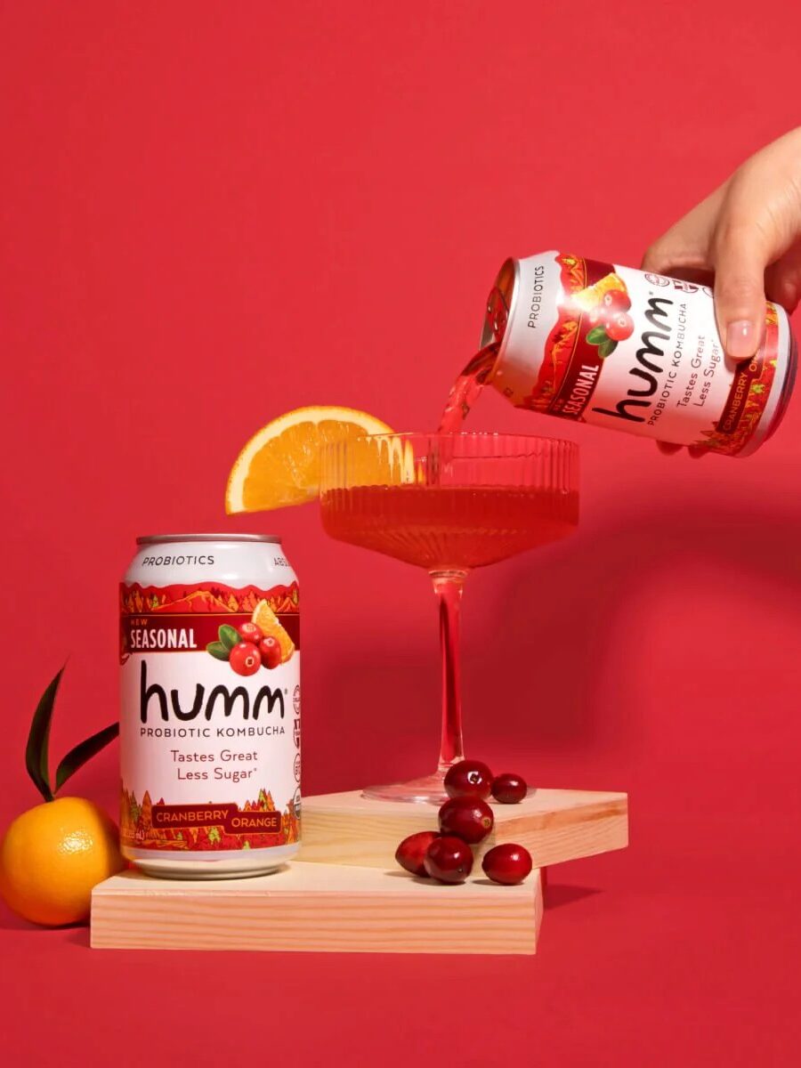 Hand pouring Humm cranberry orange kombucha into a glass on a red background, with a can, cranberries, and an orange slice nearby.