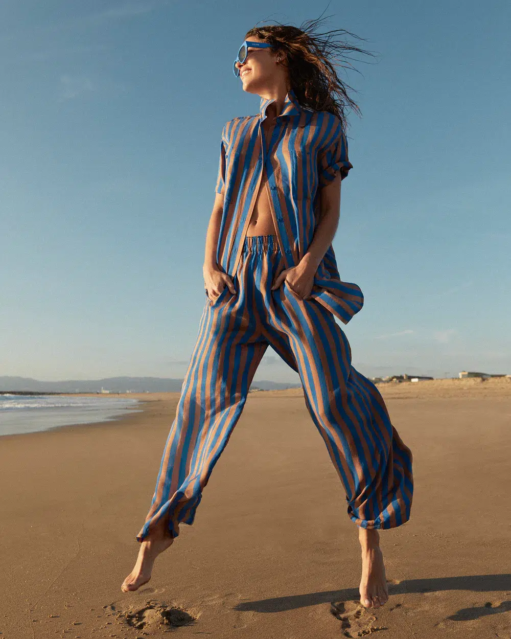 Person wearing a blue and orange striped outfit, jumping on a beach with the ocean in the background and windy hair.