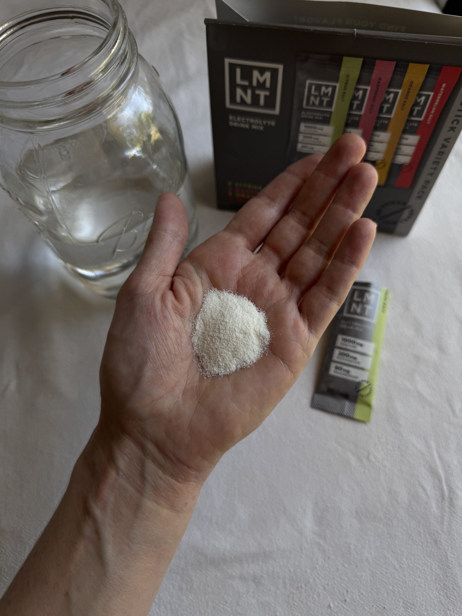 A hand holding a mound of white powder, with a glass jar of water and an open box of LMNT electrolyte drink mix packets in the background.