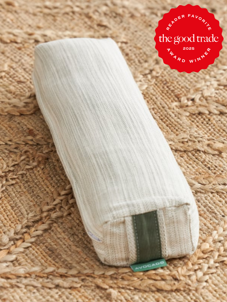 A rectangular, off-white pillow with a green tag labeled "Avocado" rests on a woven mat. A red badge in the corner reads "The Good Trade Reader Favorite 2025 Award Winner.