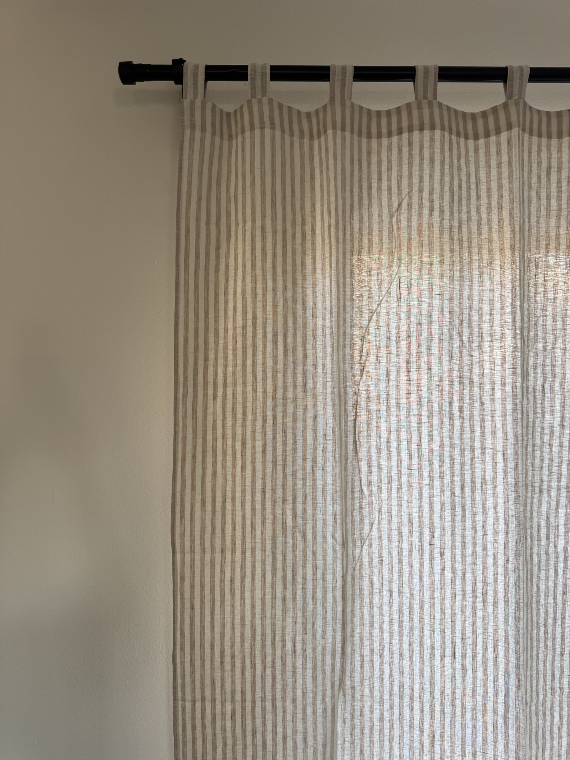 Beige and white striped curtain hangs from a black rod, partially covering a window. The wall is painted off-white. Shadows and light indicate daylight outside.