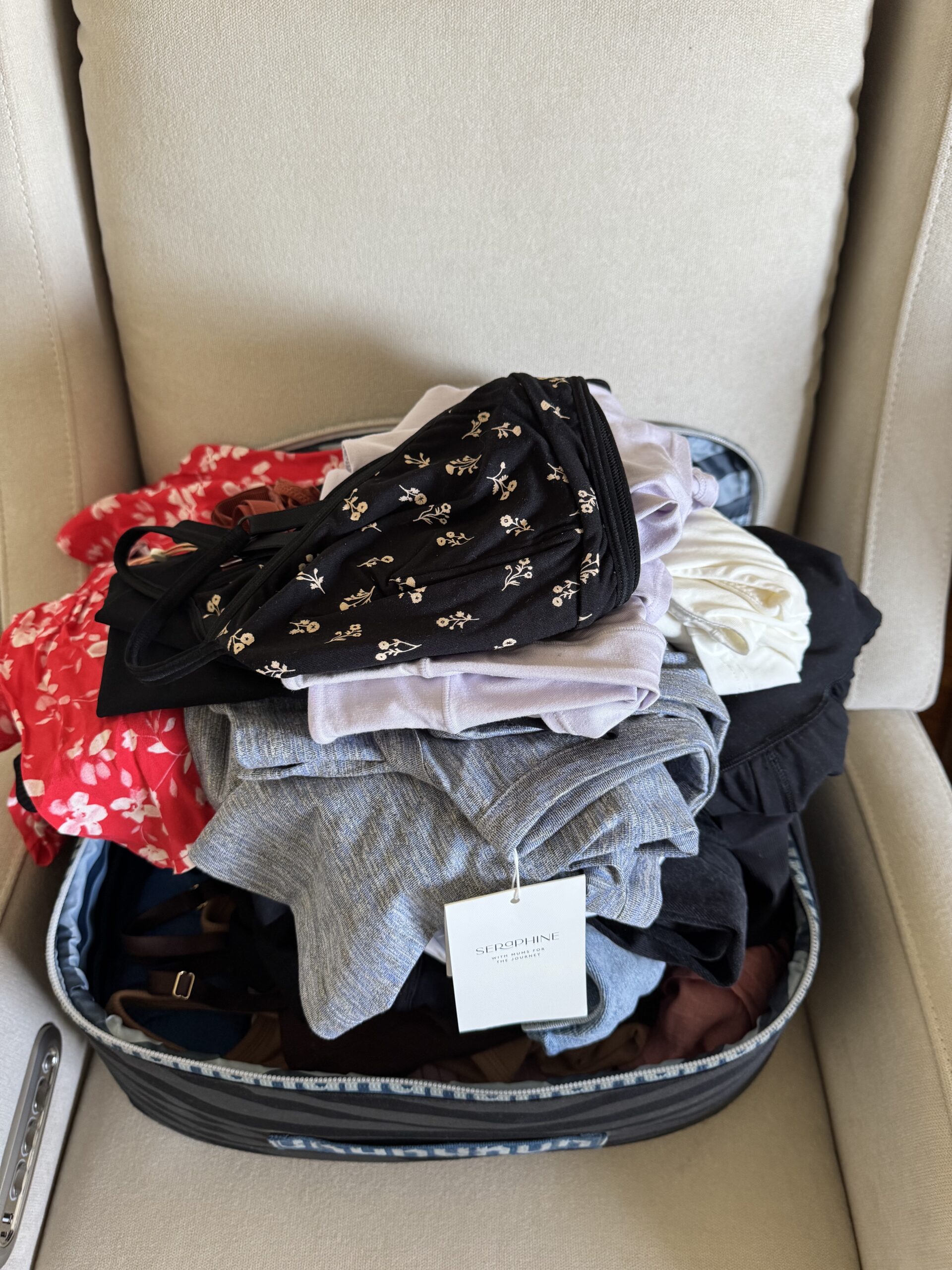 An overstuffed suitcase with various folded clothes, including floral and plain patterns, sits open on a beige armchair.