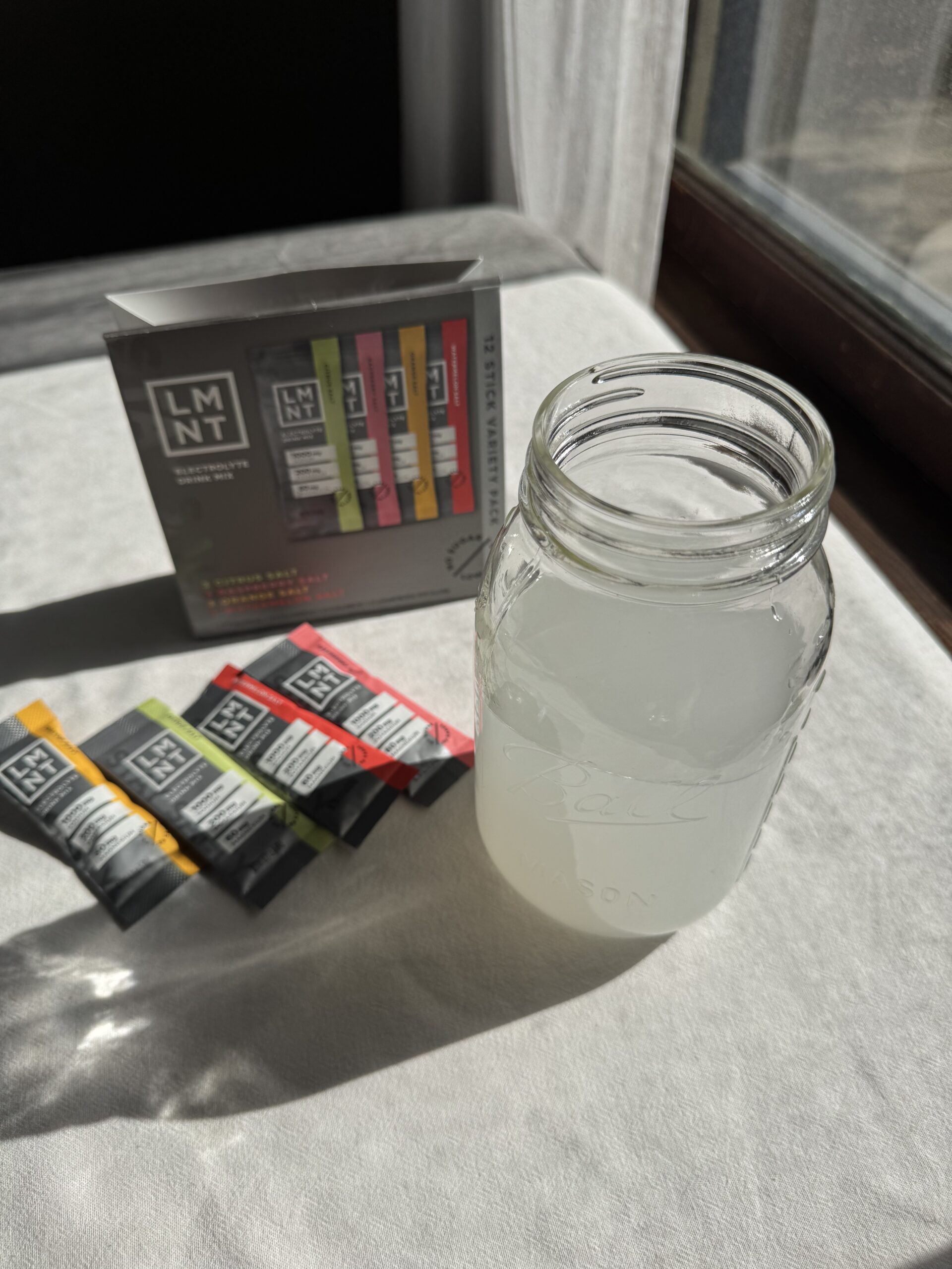 A mason jar filled with a cloudy liquid sits next to a box and four packets of LMNT electrolyte drink mix on a sunlit surface.