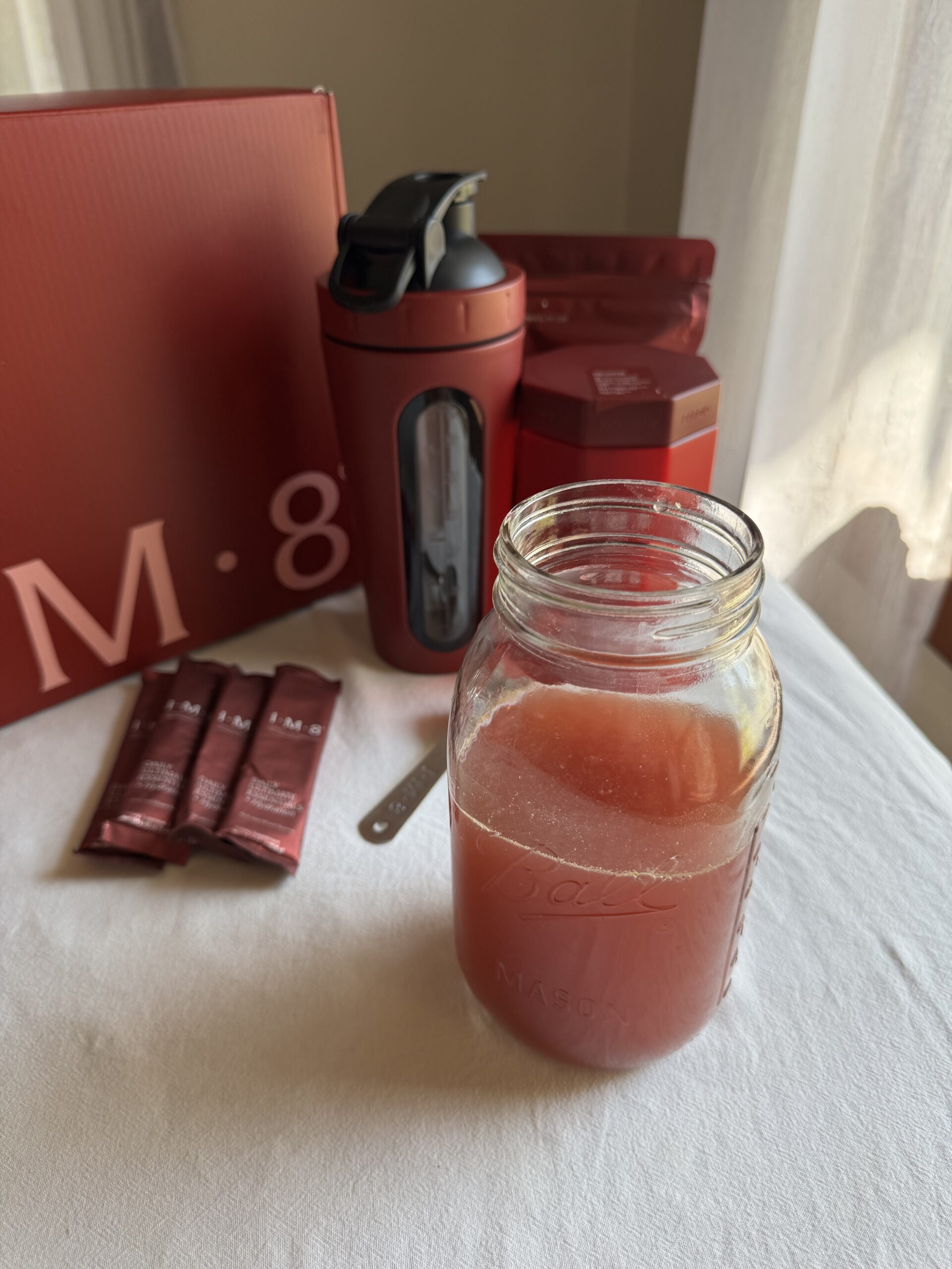A glass jar filled with a red drink sits on a table, surrounded by red product packaging, single-serve packets, a bottle, and a metal scoop.